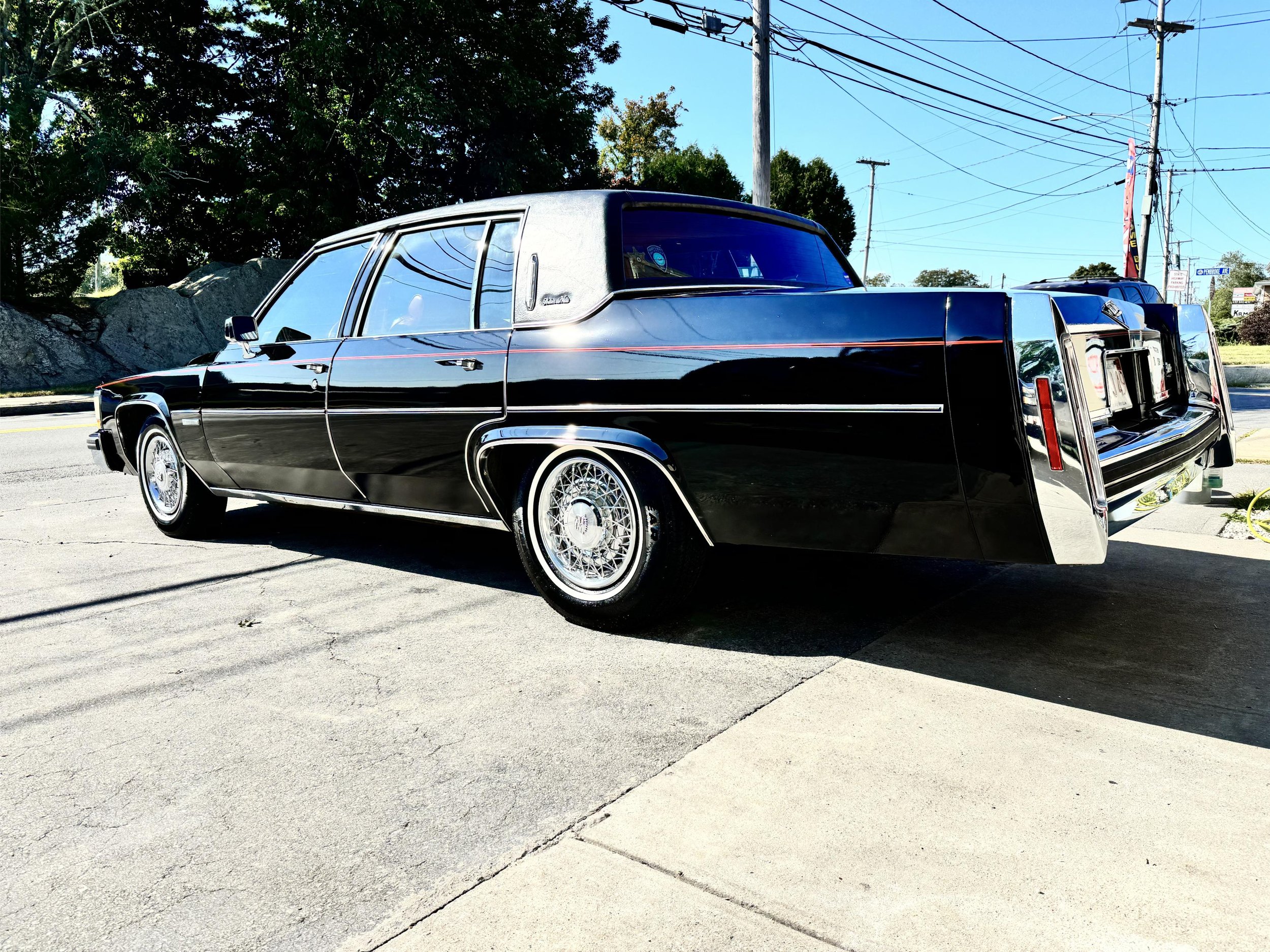 A black vintage Cadillac limousine parked on the street near a curb, with trees, power lines, and utility poles visible in the background on a bright sunny day.