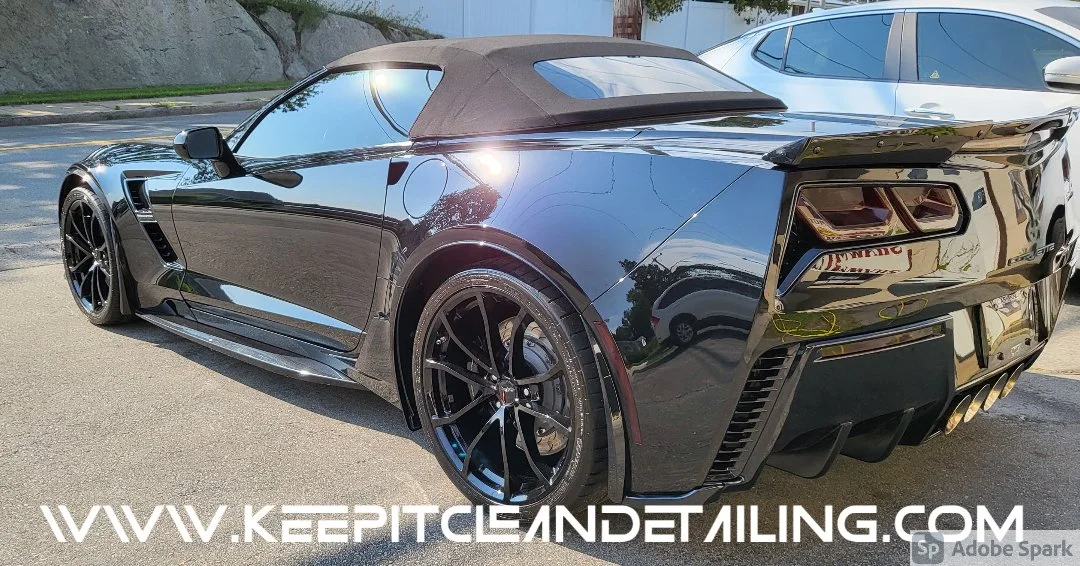 A sleek black convertible sports car with black rims parked on the street, with a silver car parked beside it and buildings and trees in the background.