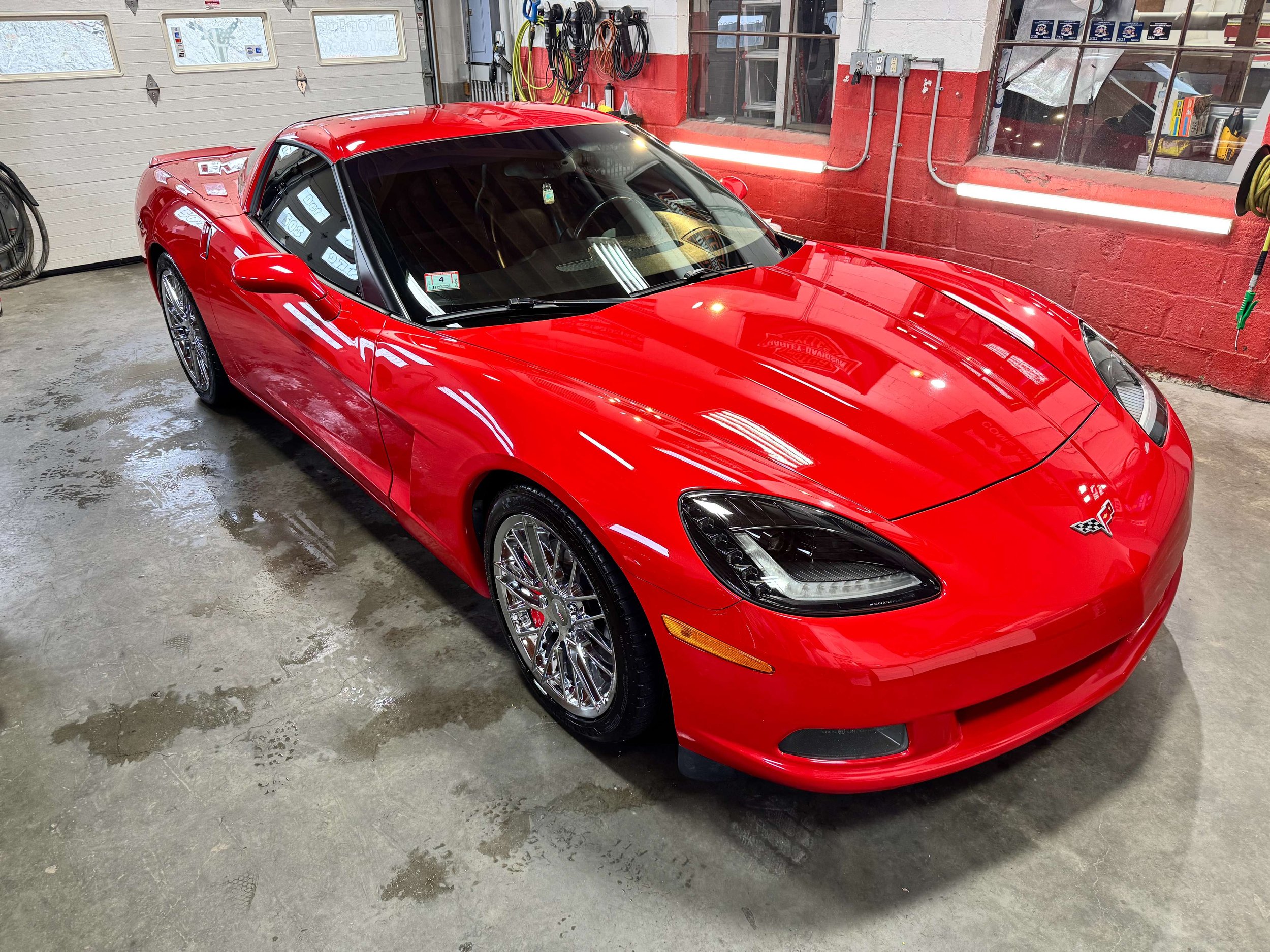A red sports car parked inside a garage with a concrete floor, red painted walls, and large windows. The garage has various hoses and tools hanging on the walls.