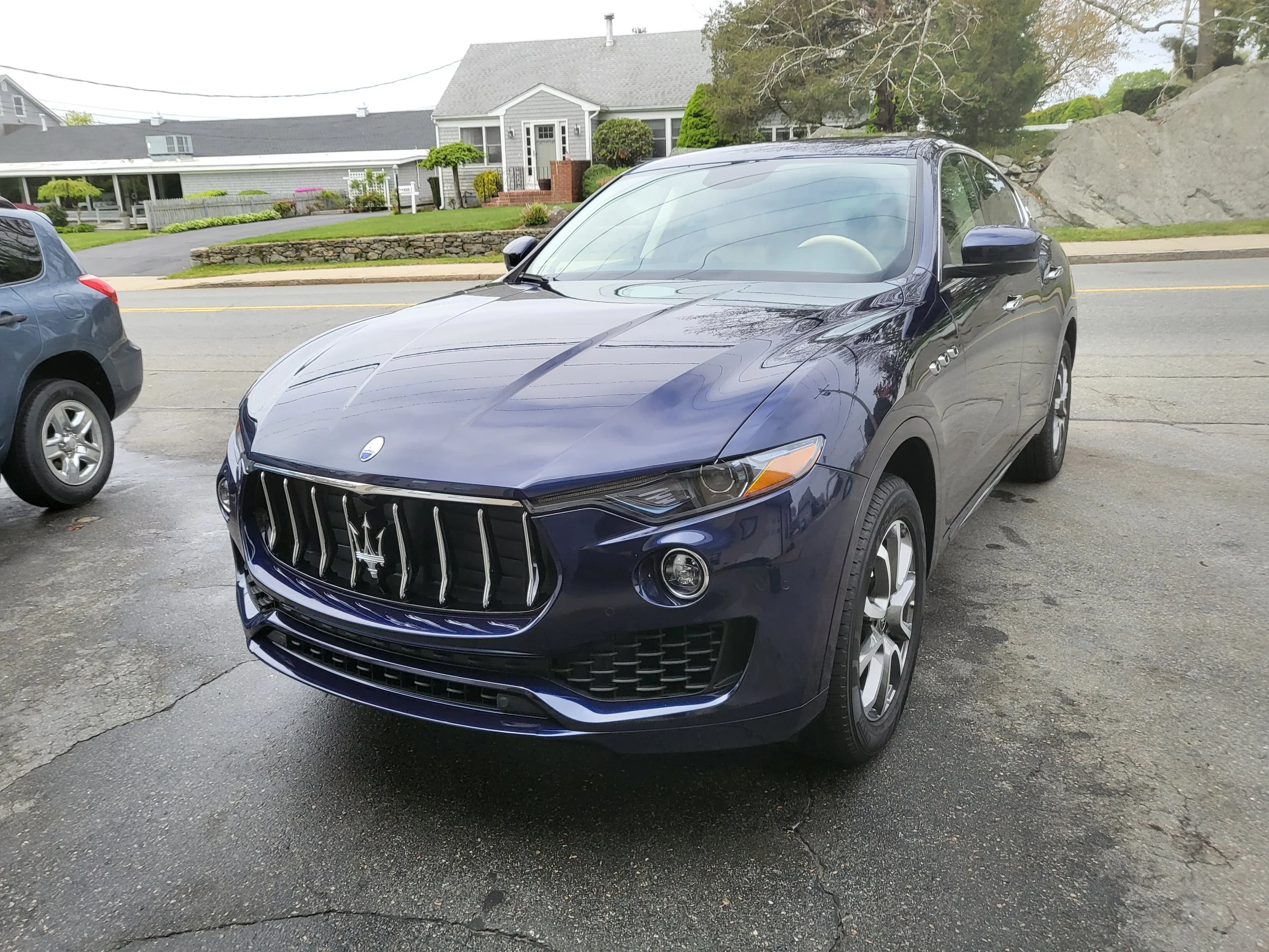 A dark blue Maserati car parked on a residential street during daytime, with a house and trees in the background.