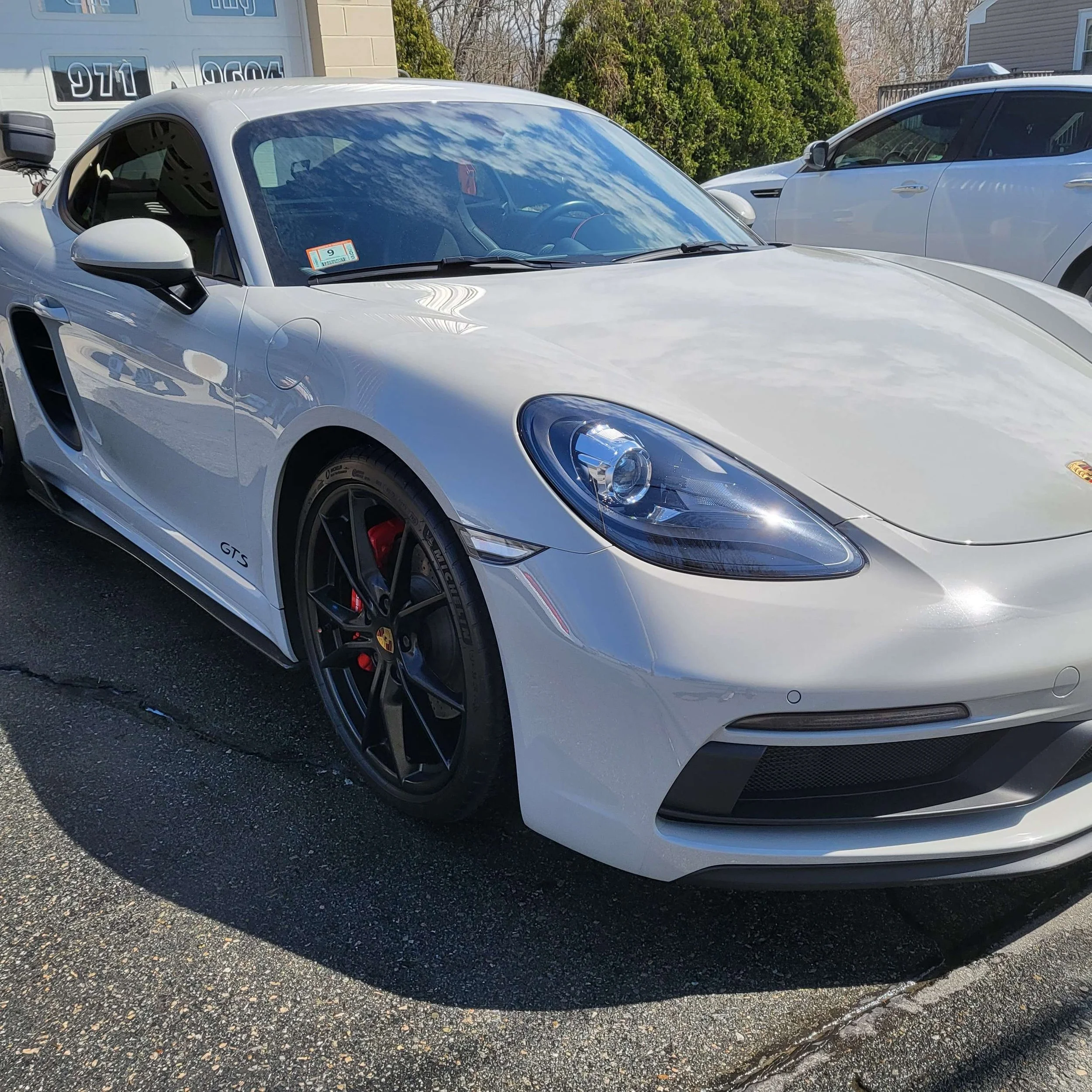 A silver Porsche sports car parked on a driveway, with another white vehicle parked next to it. The cars are in front of a garage with a white door, and there are trees and a house in the background.
