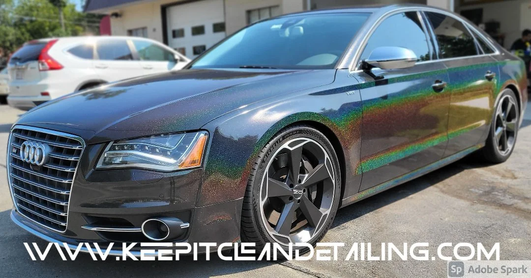 Black Audi sedan parked outdoors with metallic finish, featuring large alloy wheels and illuminated headlights.