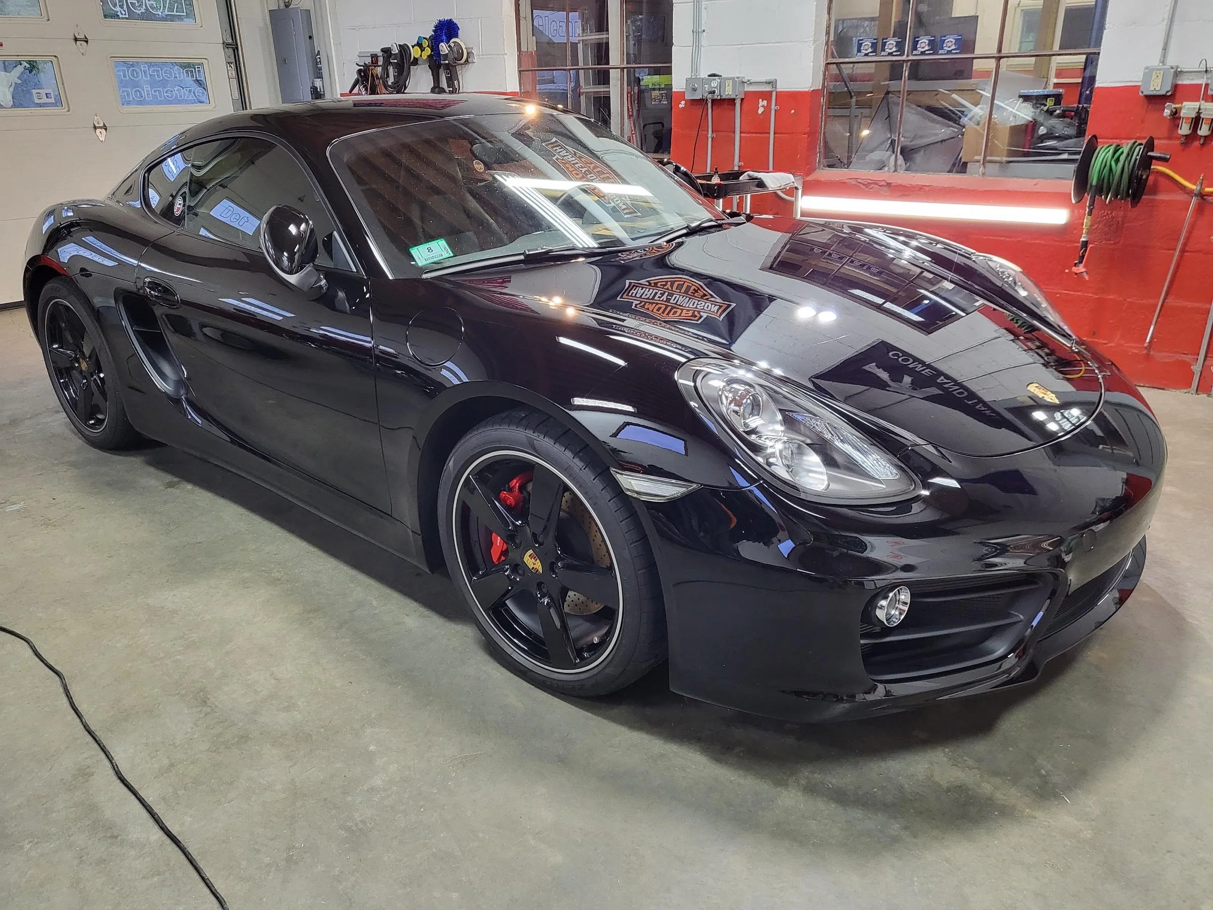 A black sports car parked inside a garage, with a red wall and various tools and hoses on the wall in the background.