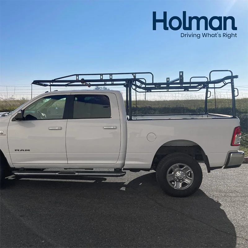 White pickup truck with a black ladder rack on top parked on the road, with a blue sky and a grassy hill in the background, and Holman logo in the top right corner.