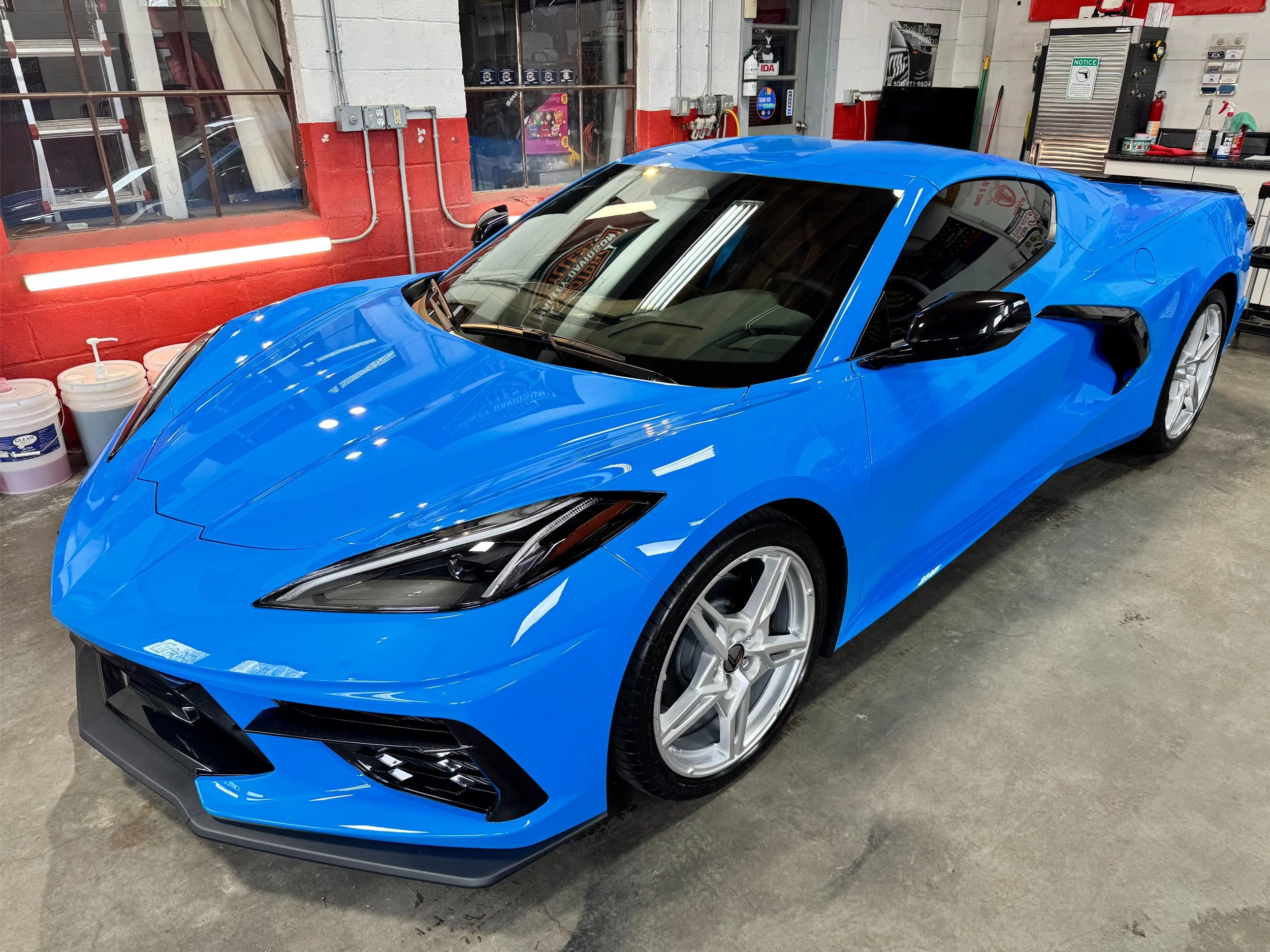 A shiny blue sports car parked inside a garage with exposed brick walls and various tools and equipment on shelves in the background.