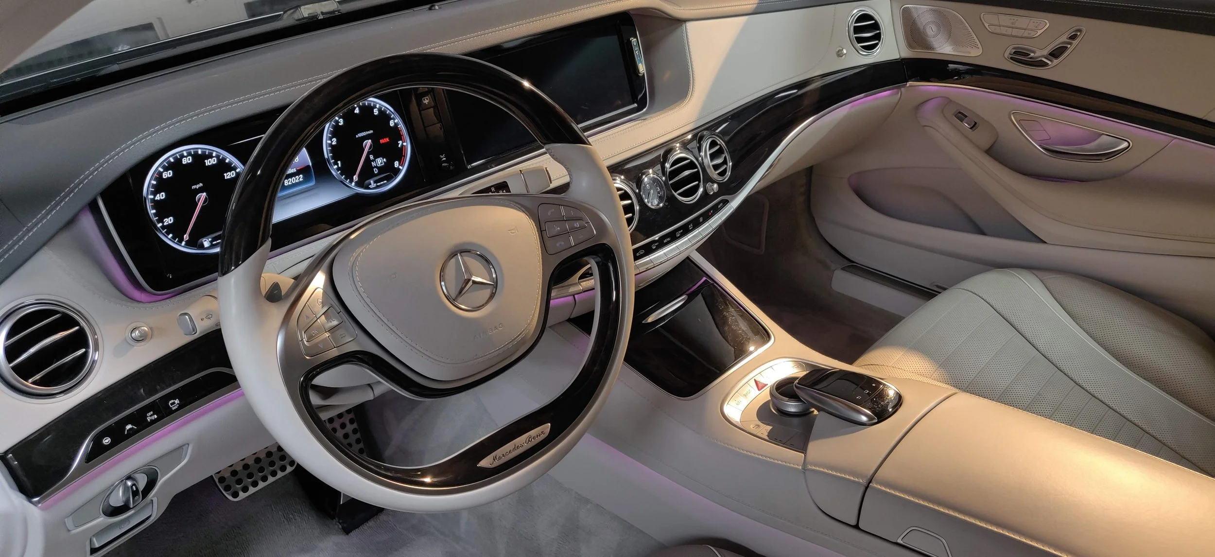 Interior of a luxury Mercedes-Benz car with light-colored leather seats, a dashboard with digital displays, and ambient purple lighting.