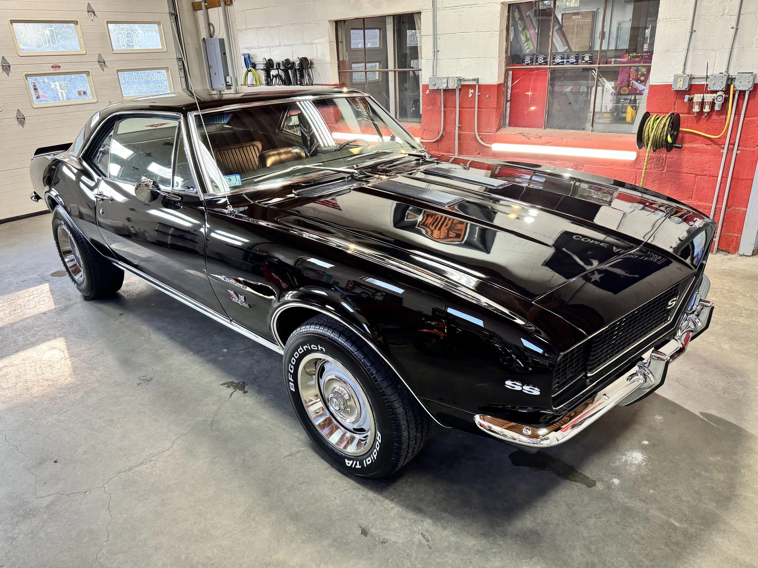 A black classic Chevrolet Corvette car parked inside a garage with a red painted wall, yellow hose reel, and various tools and electrical boxes mounted on the wall.