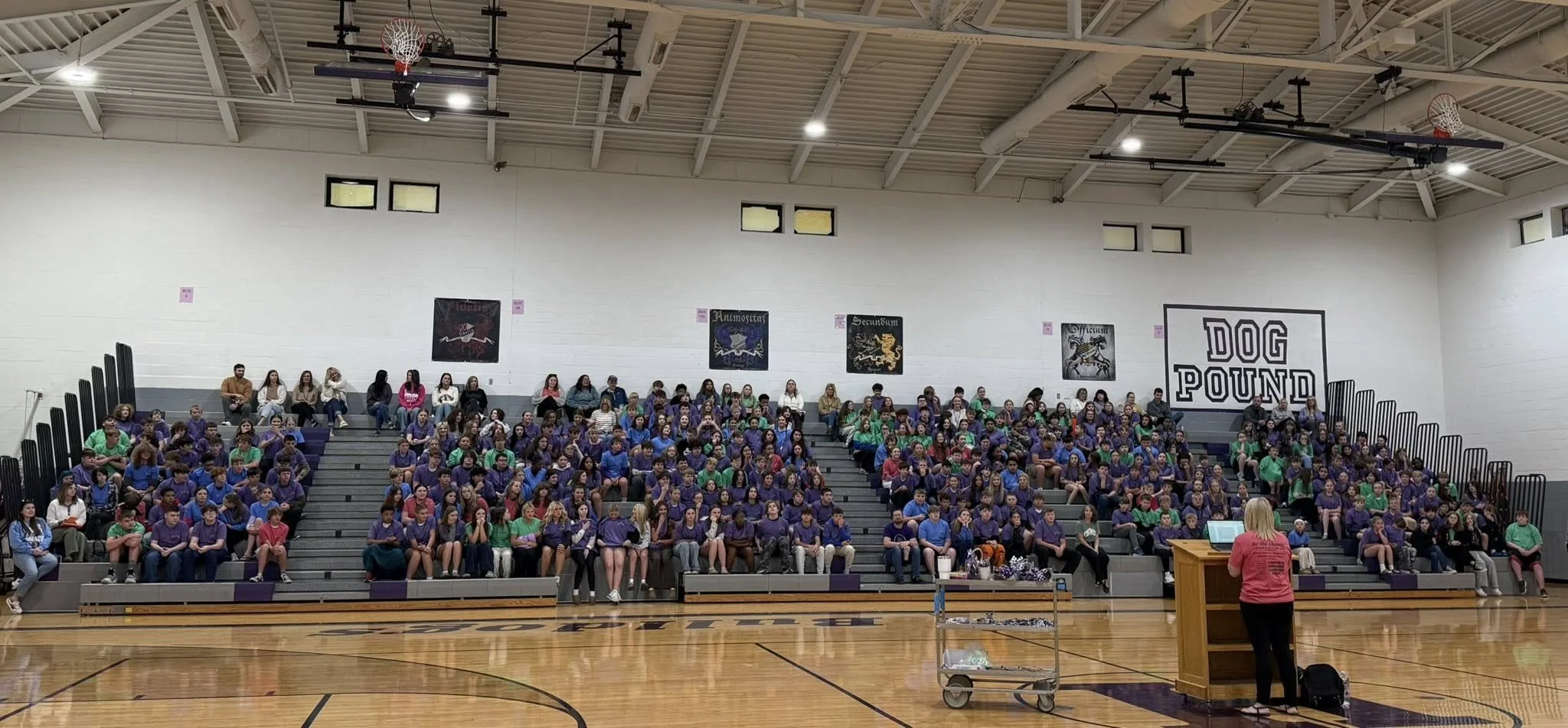 A large group of students sitting on bleachers in a school gymnasium, listening to a woman speaking at a podium.