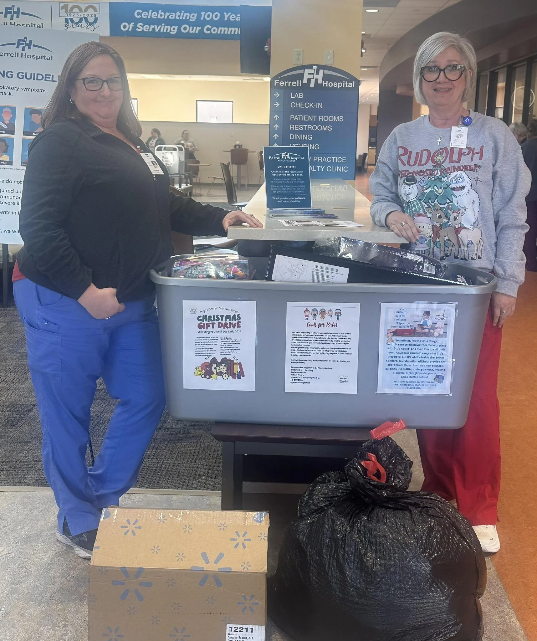 Two women standing behind a table at a donation event in a hospital lobby. The woman on the left is wearing glasses, a black jacket, and blue pants. The woman on the right is wearing glasses, a gray Rudolph Christmas sweatshirt, and red pants. The table has donation flyers, a cardboard box, and a large black trash bag. Behind them are signs and banners for Ferrell Hospital, indicating the event is a Christmas gift drive and coat drive for kids.