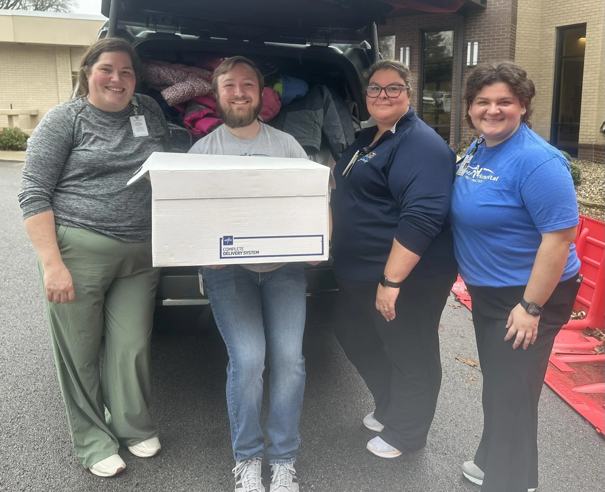 Four people standing outside at a community event, smiling, with an open trunk of a vehicle filled with bags and boxes behind them.