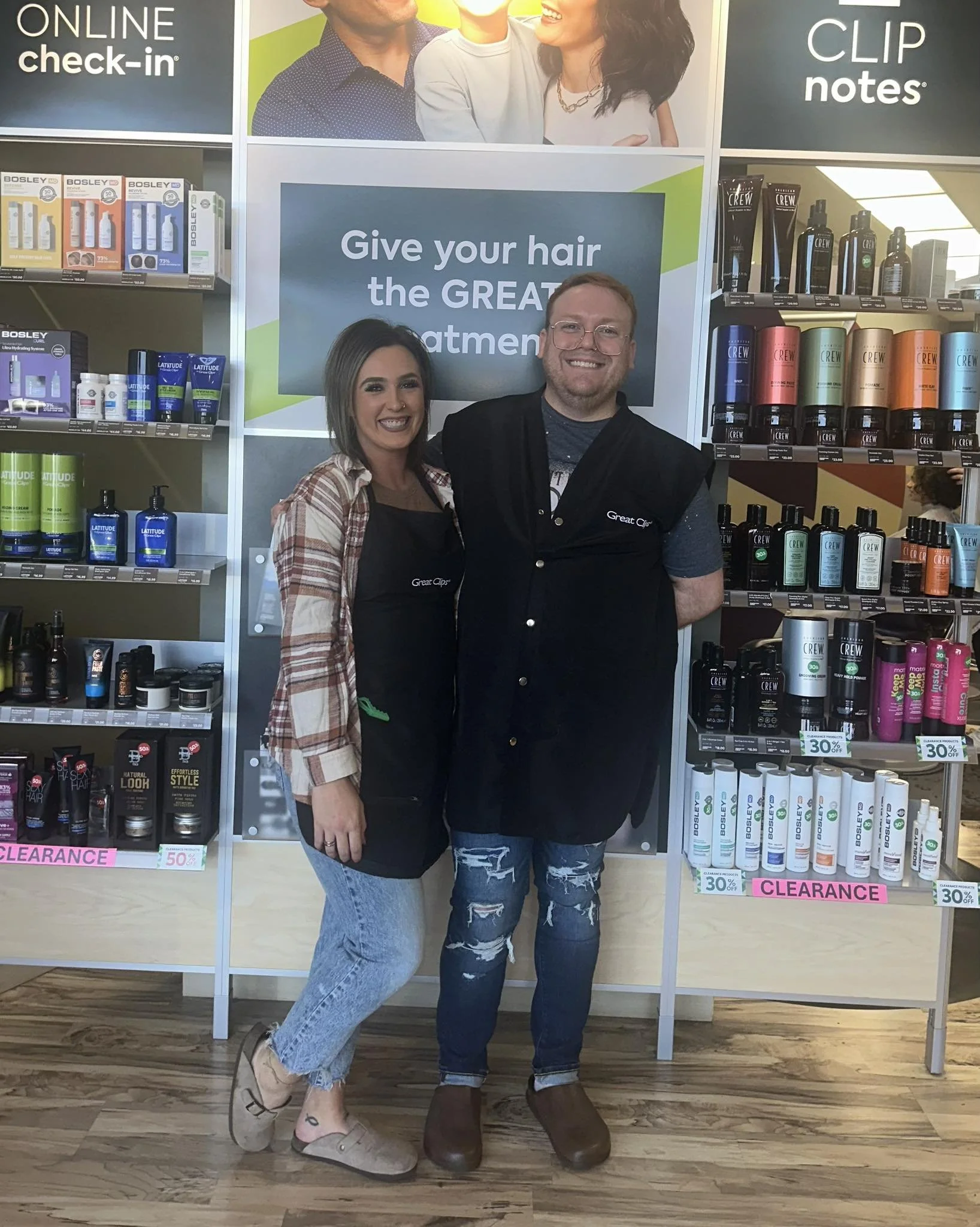 Two women and a man smiling and standing close to each other in a store aisle. The woman on the left has dark hair, is wearing a plaid shirt and ripped jeans, and has tattoos on her ankle. The man in the middle has light hair, glasses, and is wearing a dark vest over a shirt with a logo. The woman on the right has dark hair, is wearing a gray shirt, and ripped jeans. They are surrounded by shelves of hair care products and large signs, including one that says 'Give your hair the GREAT treatment.'