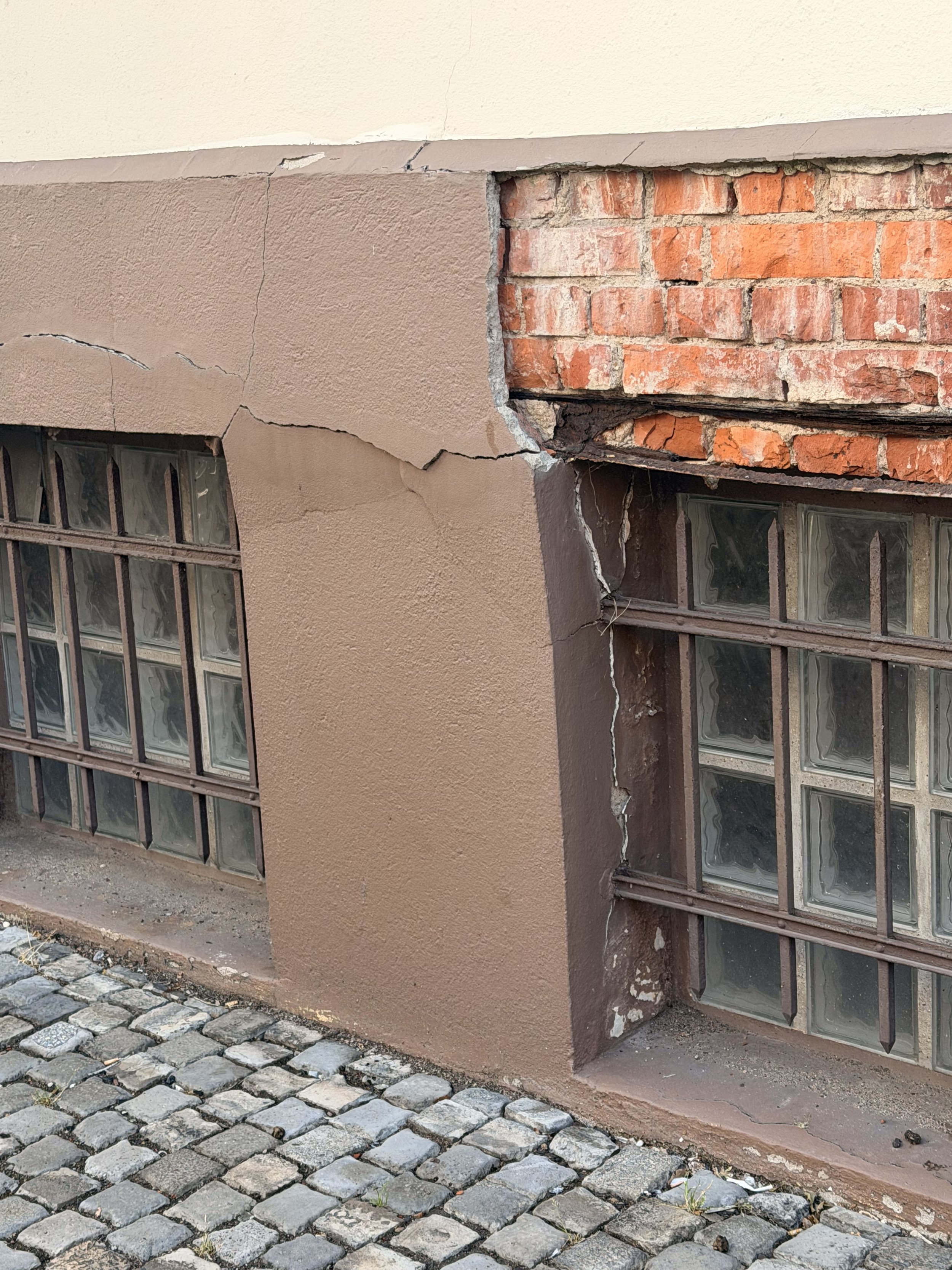 Ein Gebäude mit beschädigter Fassade, Ziegelsteinwand, Rissen in der Wand und zerbrochenem Mauerwerk. Die Fenster sind mit Metallgittern gesichert, auf dem Bürgersteig liegen Kopfsteinpflaster.