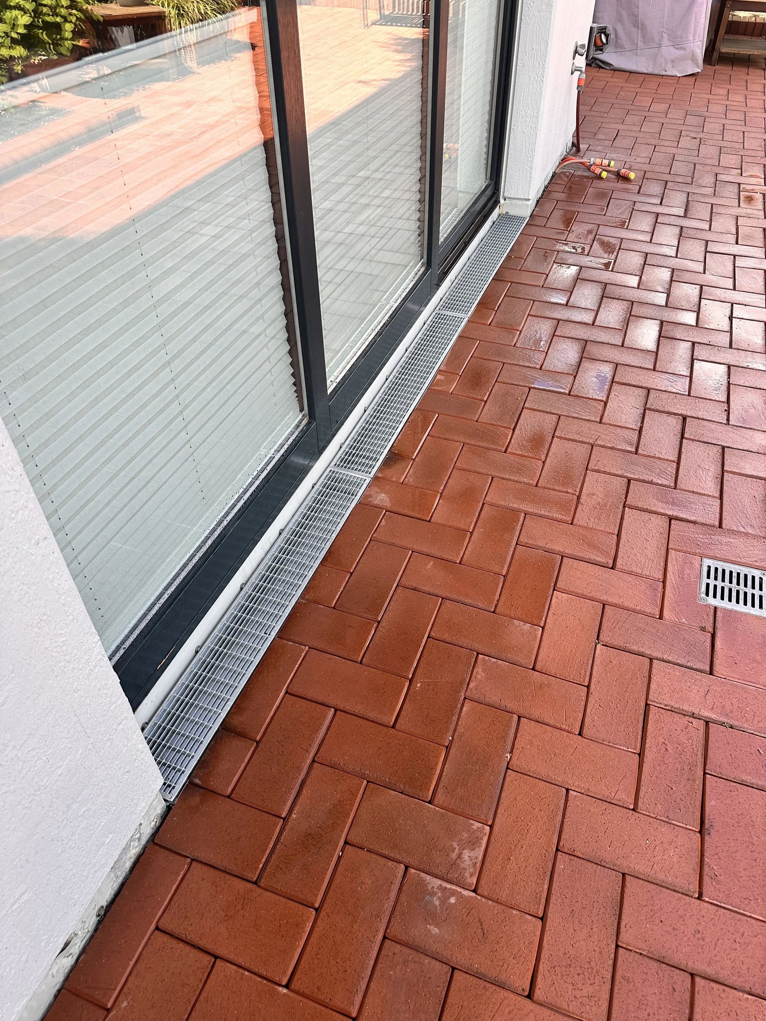 Rote gepflasterte Terrasse mit einem Abflussgitter, einer Regenrinne und einer Glasschiebetür, äußerlich, nach Regen