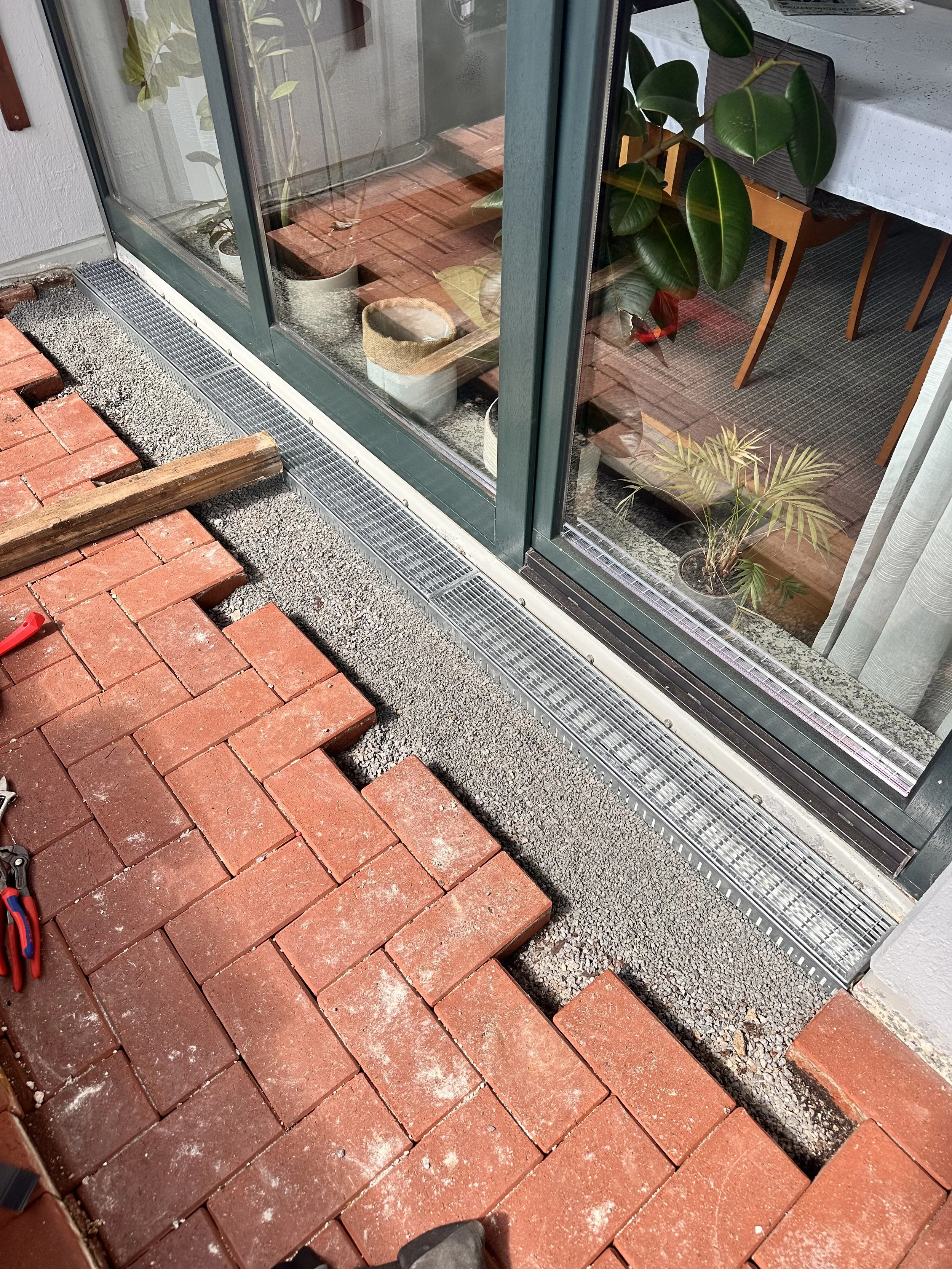 Boden im Außenbereich mit roten Ziegelsteinen und Sand, ein Teil der Fensterfront eines Hauses mit Pflanzen im Inneren.