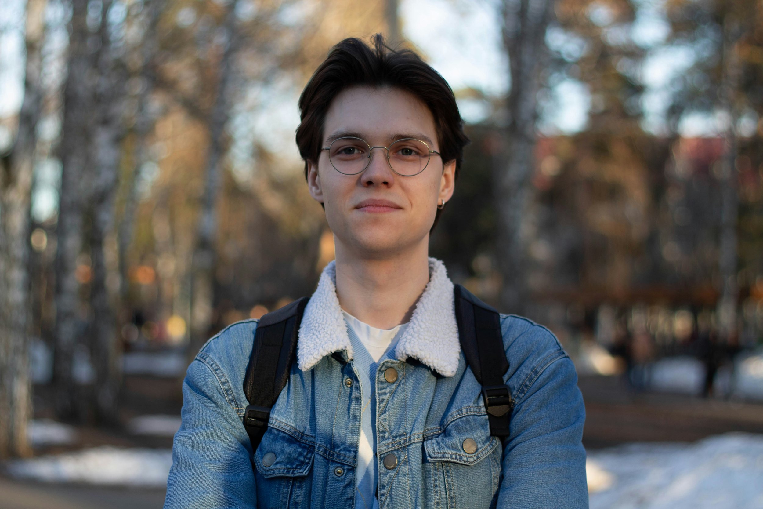 A young man with short dark hair, glasses, and earrings, wearing a denim jacket with a white fleece collar and a backpack, standing outdoors during autumn with blurred trees in the background.