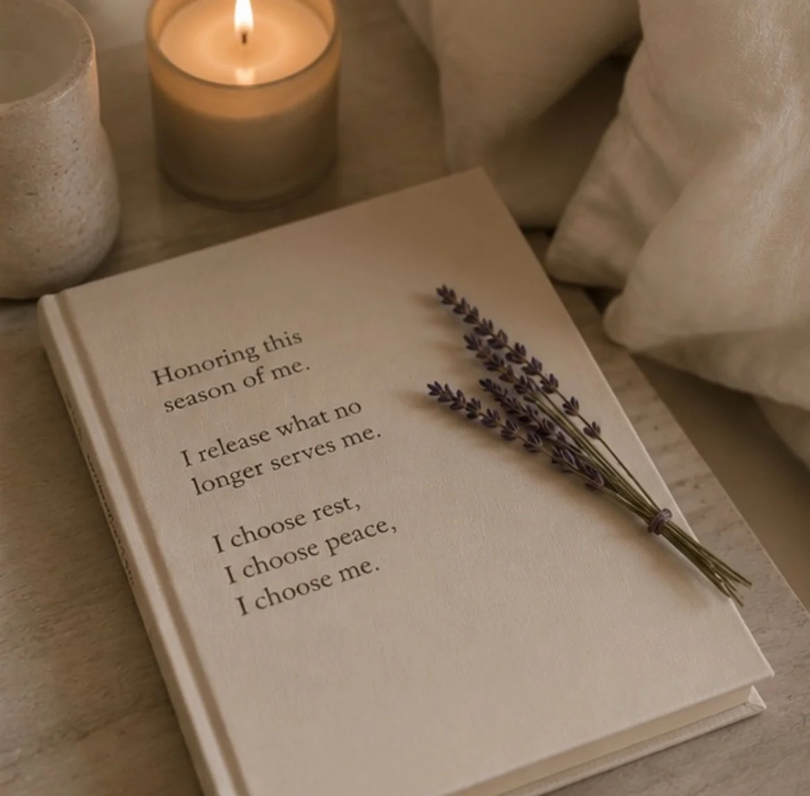 A beige-colored book with a poem on the cover, laying on a table alongside a sprig of lavender. Two candles are lit in the background, and part of a beige cloth and a textured beige vase are also visible.