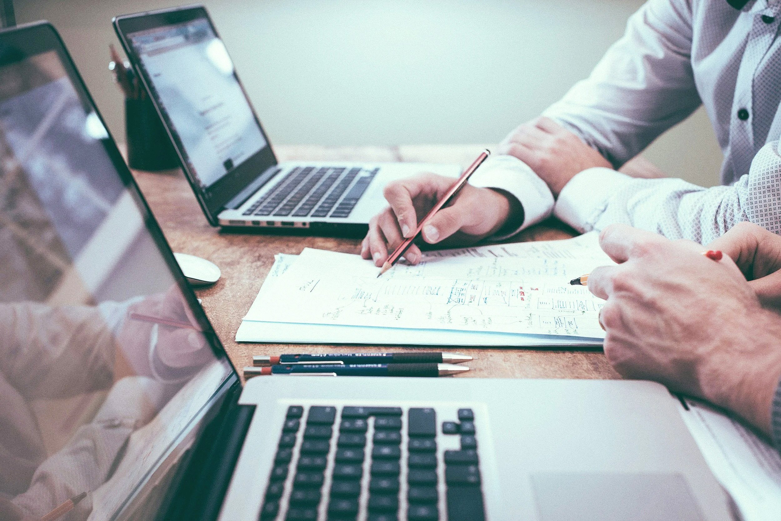 People working at a table with laptops, notebooks, pens, and documents during a meeting or collaboration.