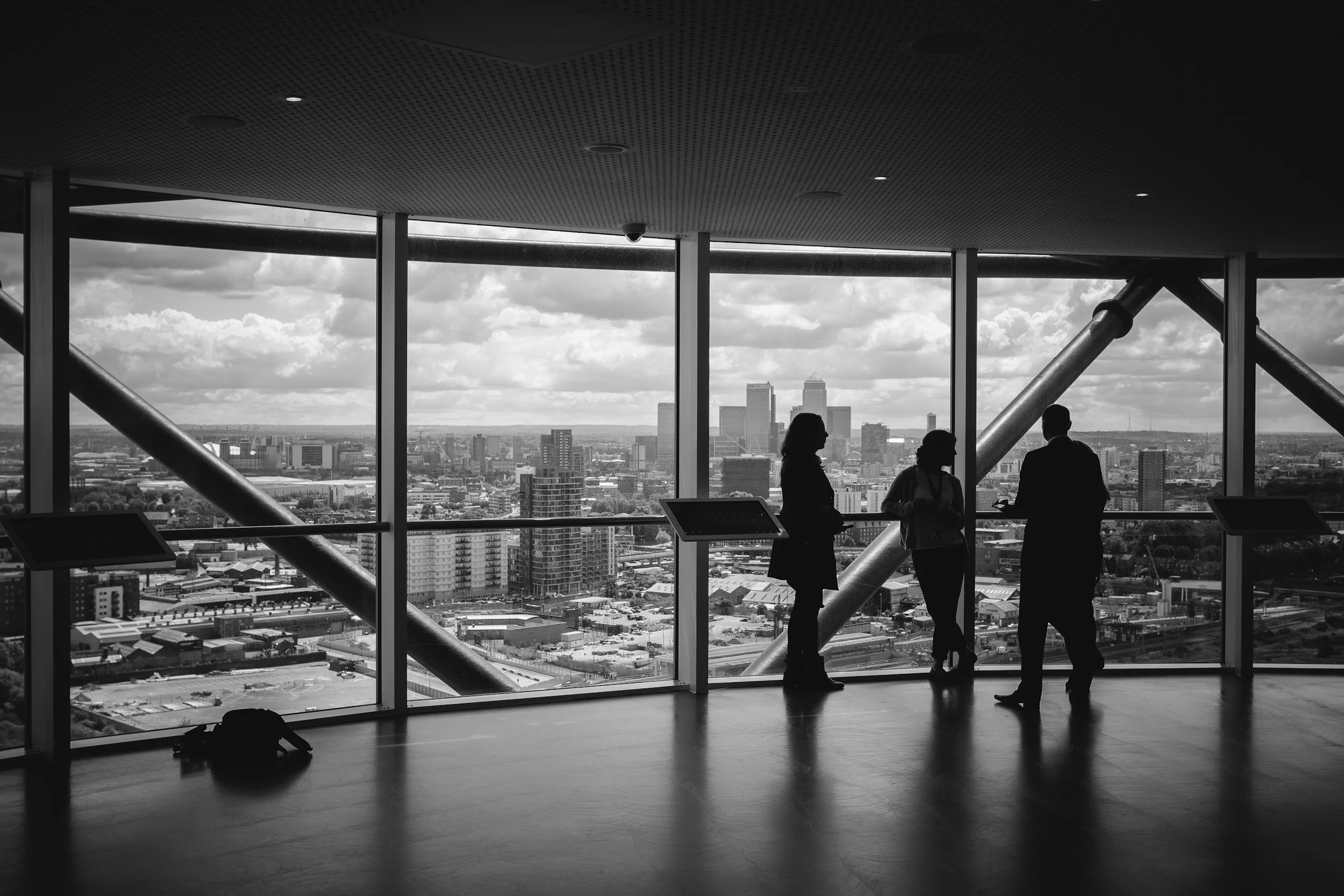 Silhouettes of three people talking inside a high-rise building with large windows, overlooking a city skyline with tall buildings and cloudy sky.