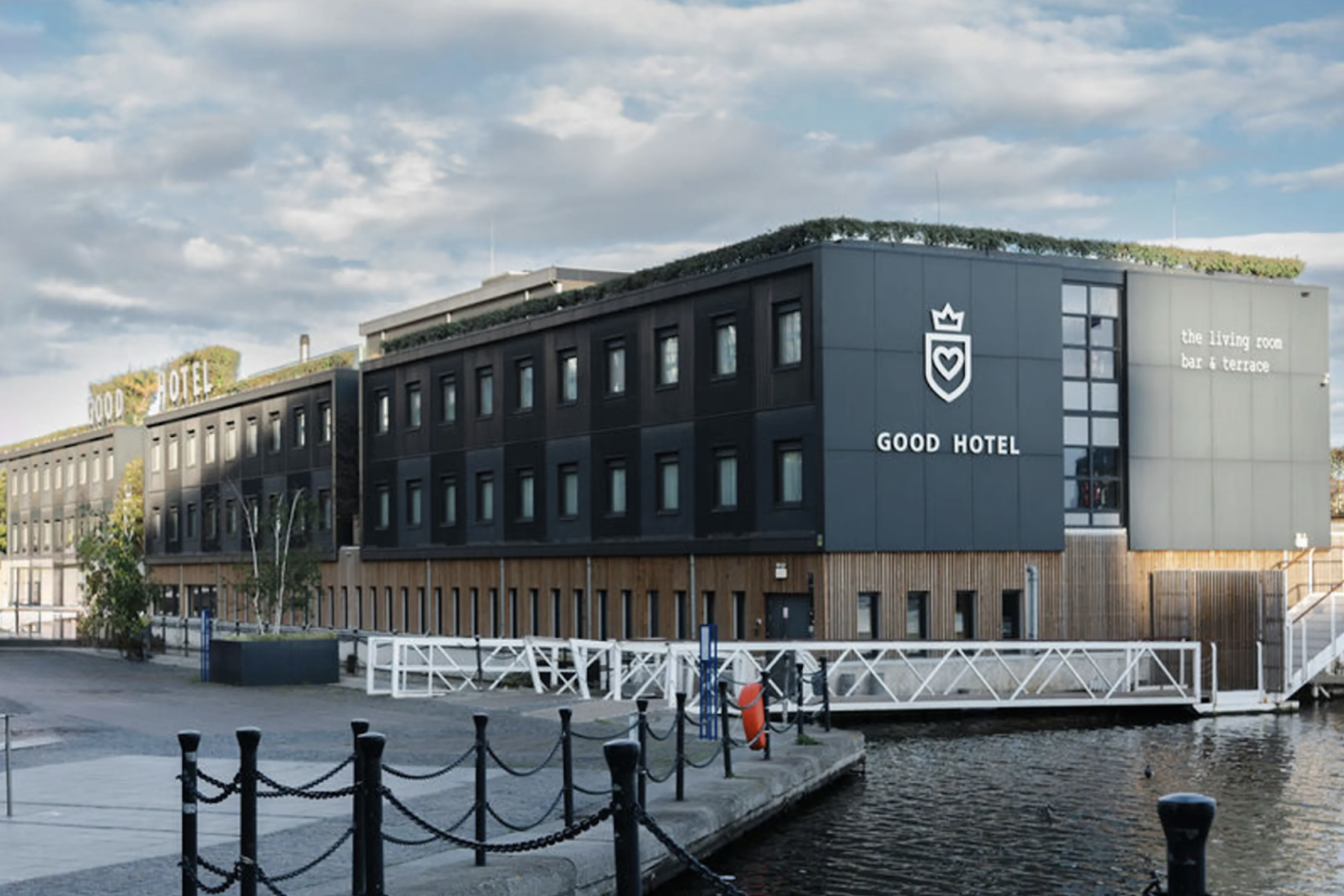 Modern building labeled 'Good Hotel' with signage indicating it has a bar and terrace, situated along a waterway with a small dock and a walking path in the foreground, under a partly cloudy sky.
