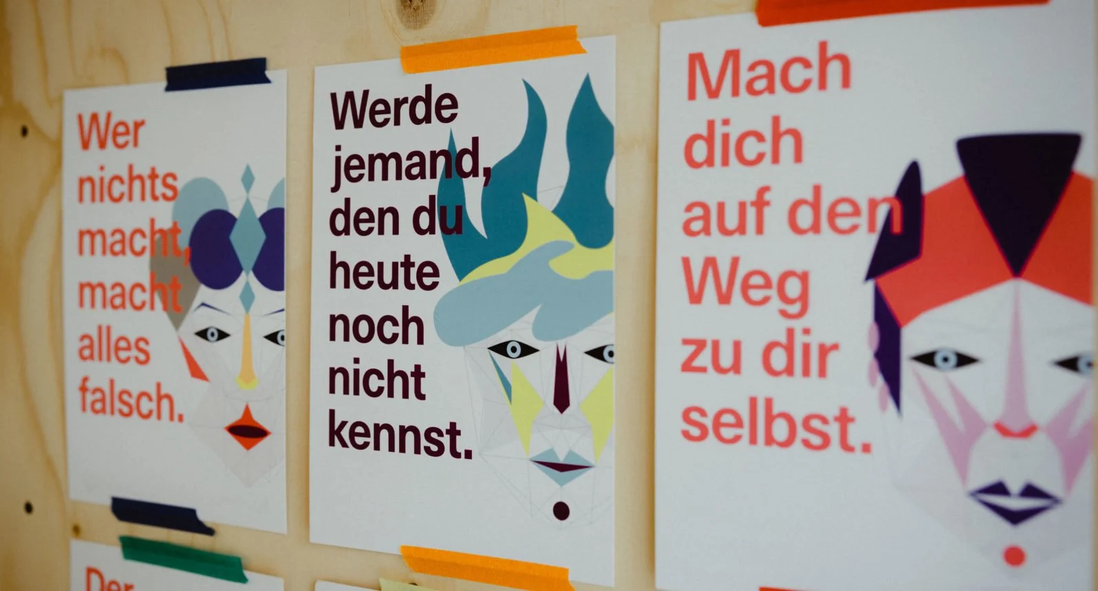 Three motivational posters with abstract faces and German text on a wooden wall, colorful tape at the top and bottom of each poster.