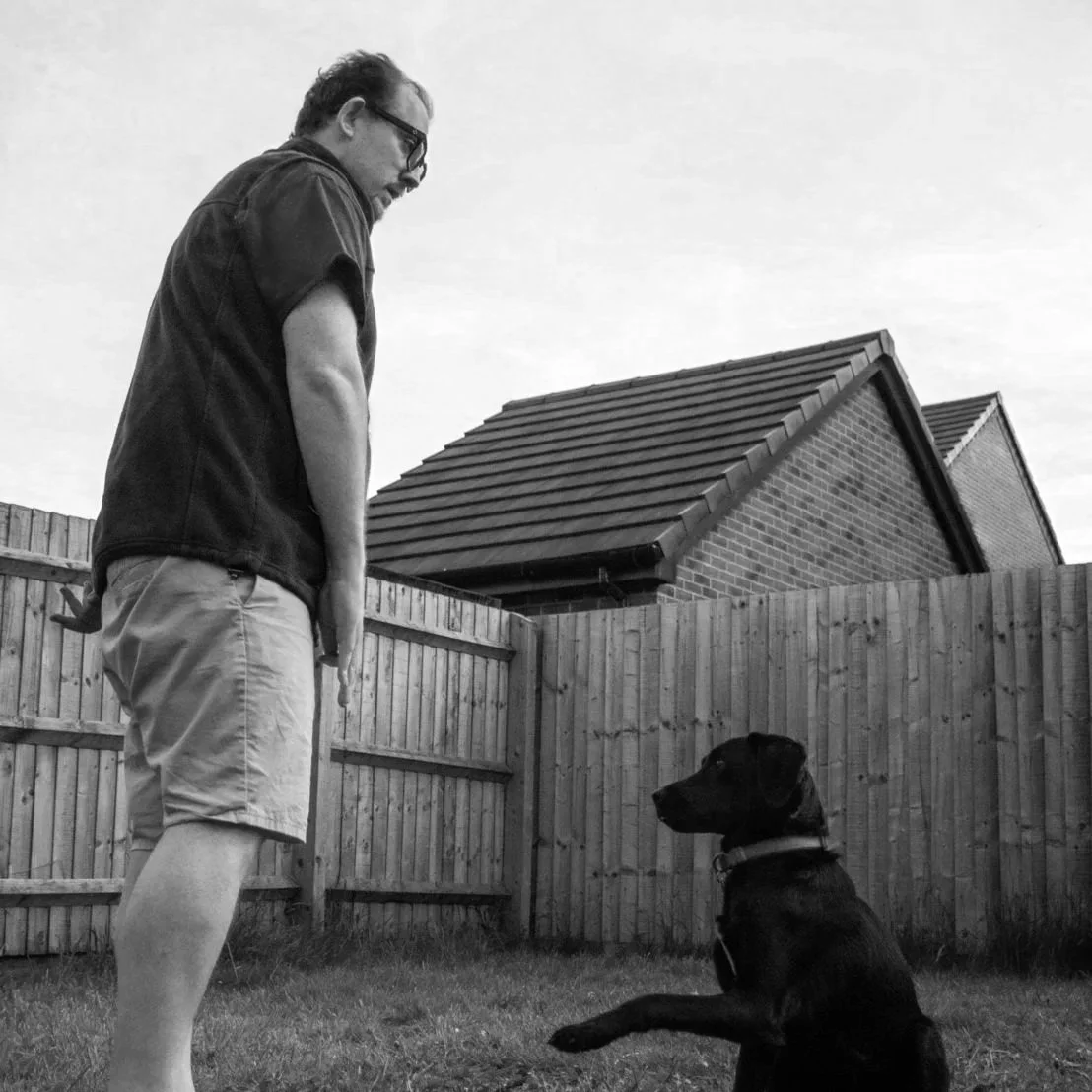 A man with glasses and a beard stands in front of a black dog sitting on grass in a backyard with a wooden fence, looking at the dog.
