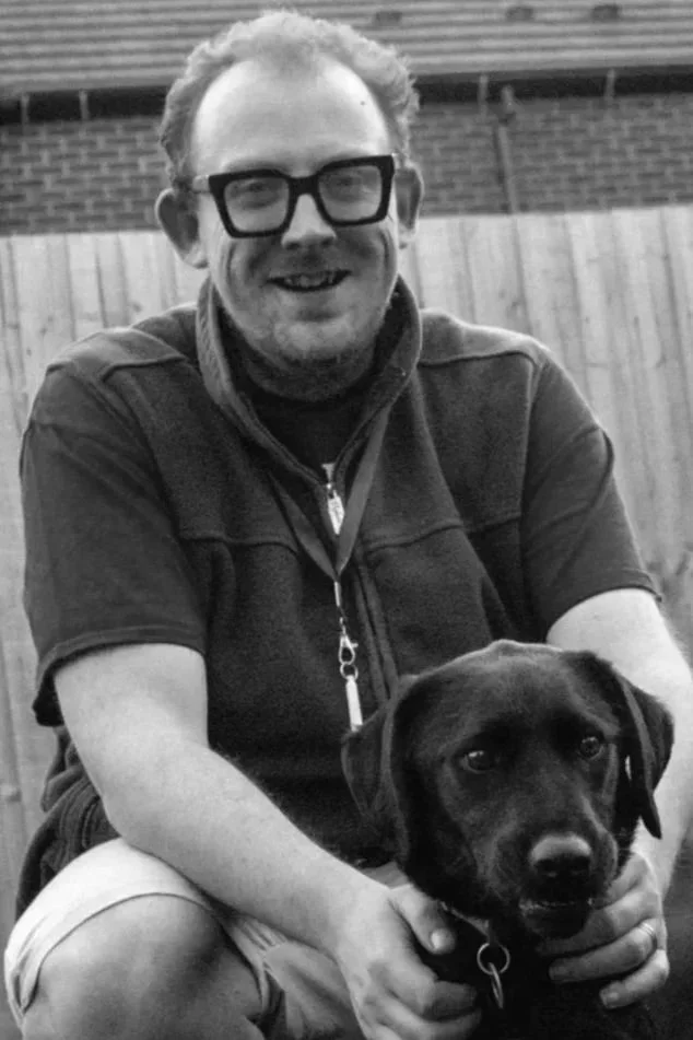 A smiling man with glasses seated outdoors with a black dog, both looking at the camera, in a backyard setting with a wooden fence.