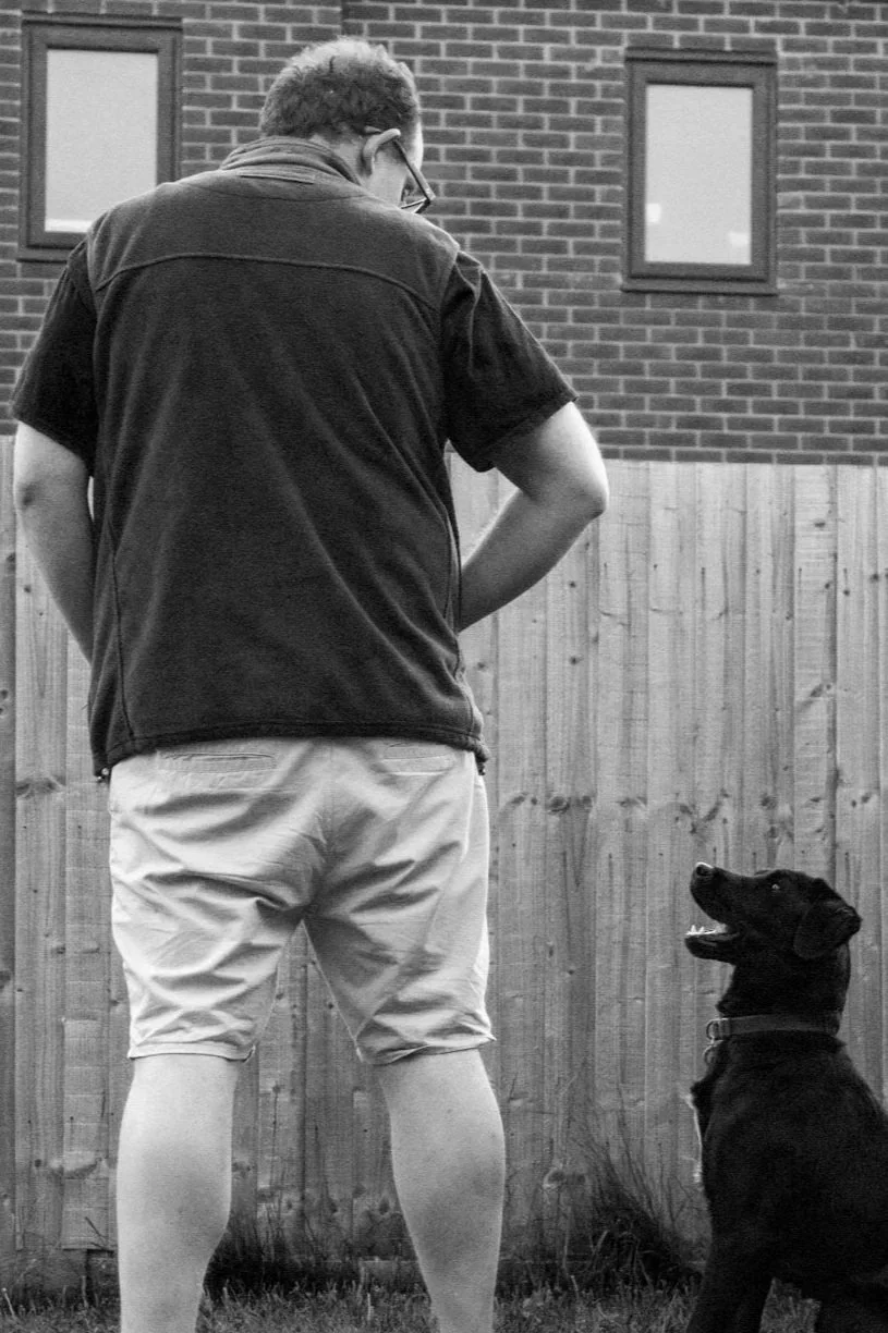 A man in shorts and a vest standing outdoors with a dog sitting and looking up at him against a wooden fence, with a brick house and two windows in the background.
