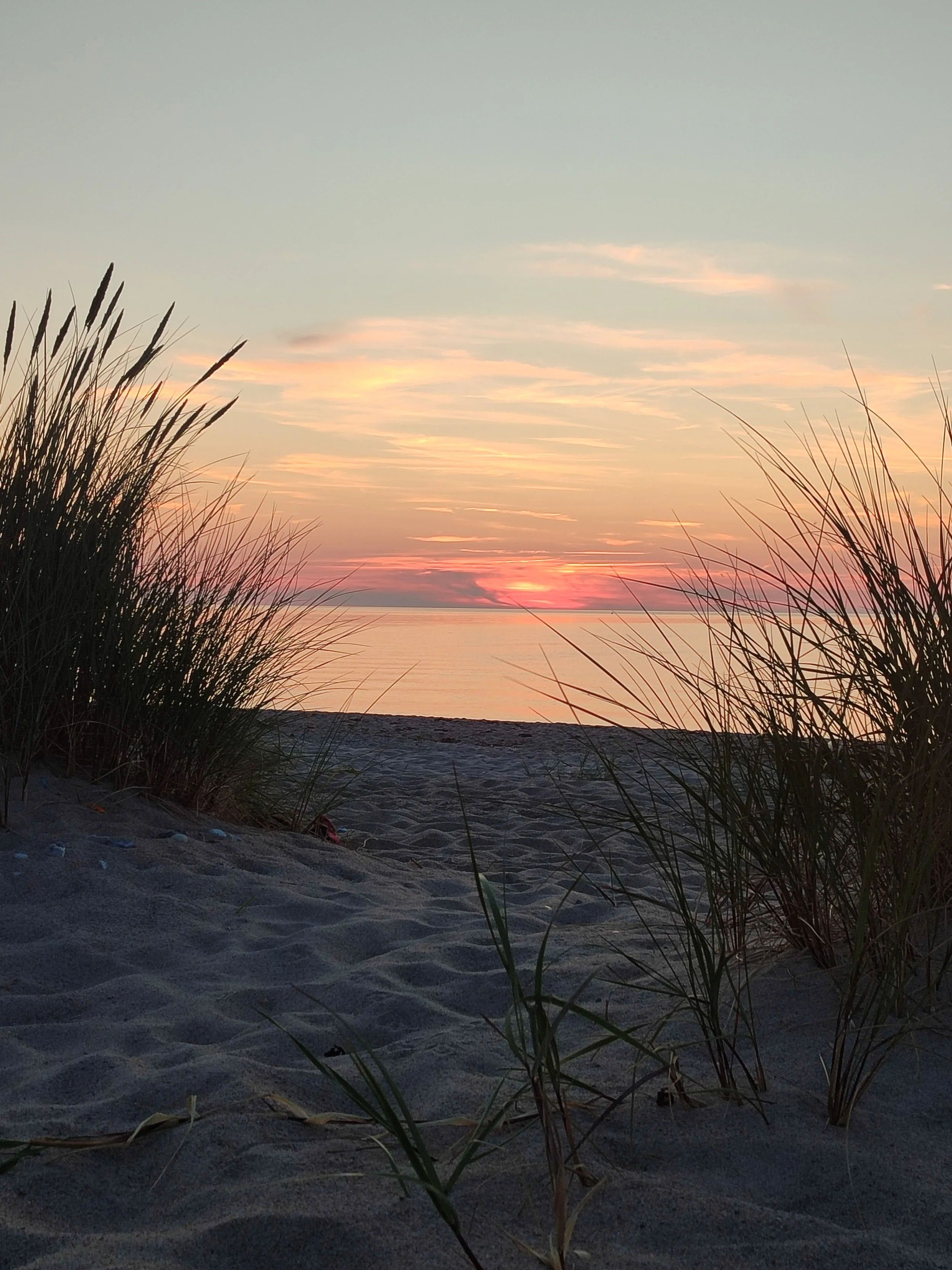 Sonnenuntergang am Strand mit Sand, Gräsern und Wasser im Hintergrund