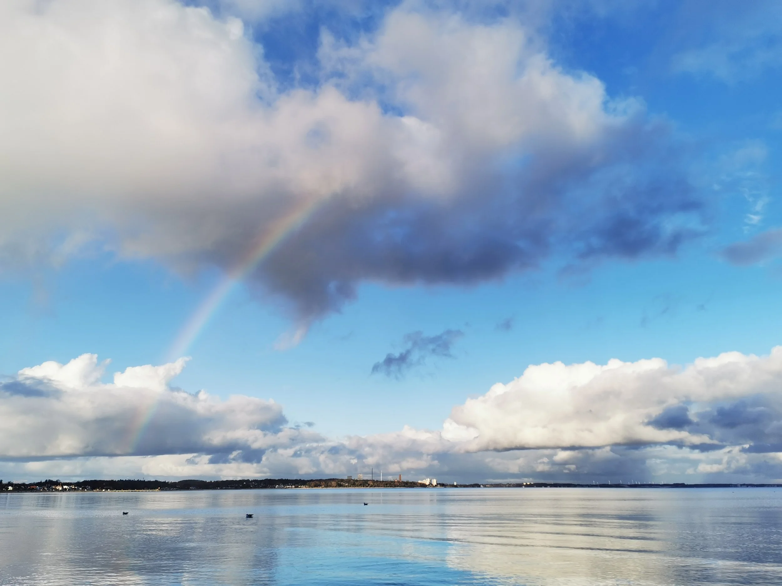 Himmel mit Wolken, Regenbogen und Wasser im Vordergrund, Stadt im Hintergrund.