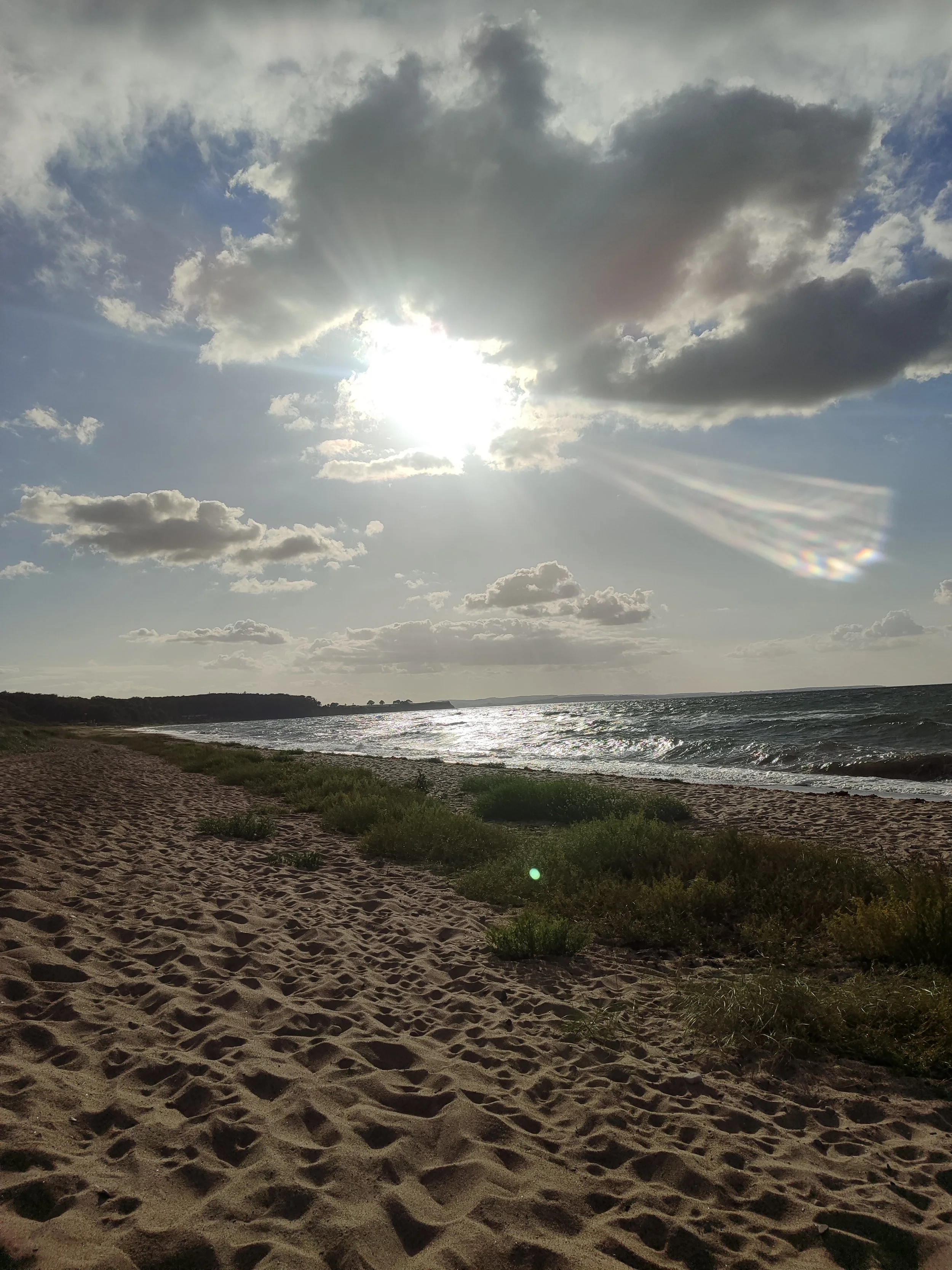 Strand mit Sand, Wasser und Wolken am Himmel, Sonne hinter Wolken sichtbar