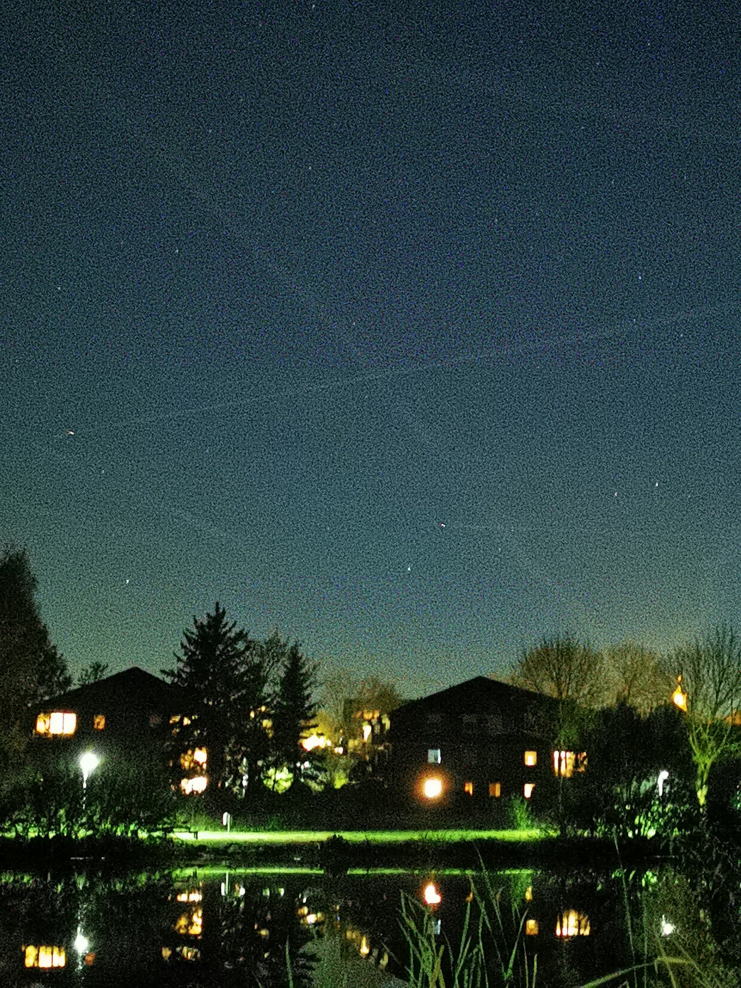 Sternenklarer Himmel über einem ruhigen See mit reflektierenden Lichtern und Silhouetten von Bäumen und Häusern im Vordergrund.