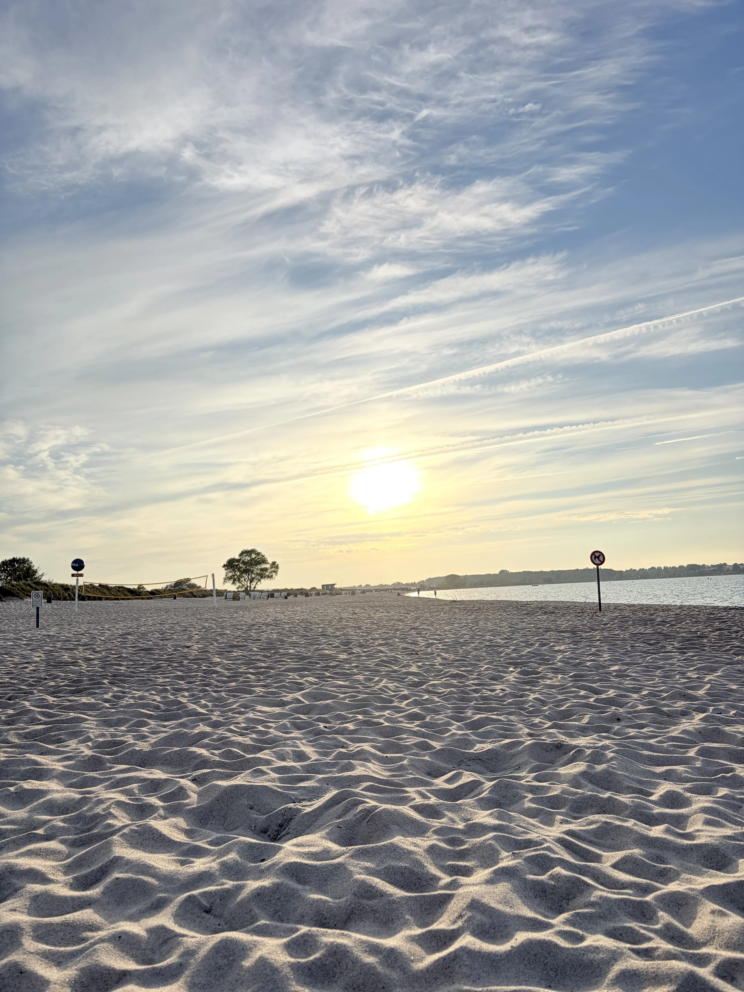 Sonnenuntergang am Strand mit sandigem Ufer, Bäumen im Hintergrund, wenige Leute und Schilder am Strand zu sehen.
