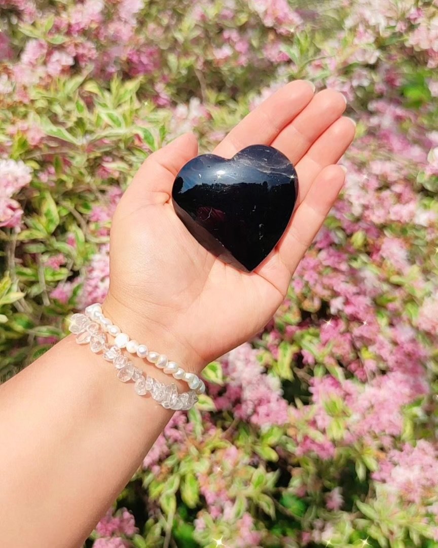 Eine Hand hält einen schwarzen, herzförmigen Stein vor einer blühenden pinken und grünen Pflanze, getragen von einer Person, die eine Perlen- und eine Kristallarmband trägt.