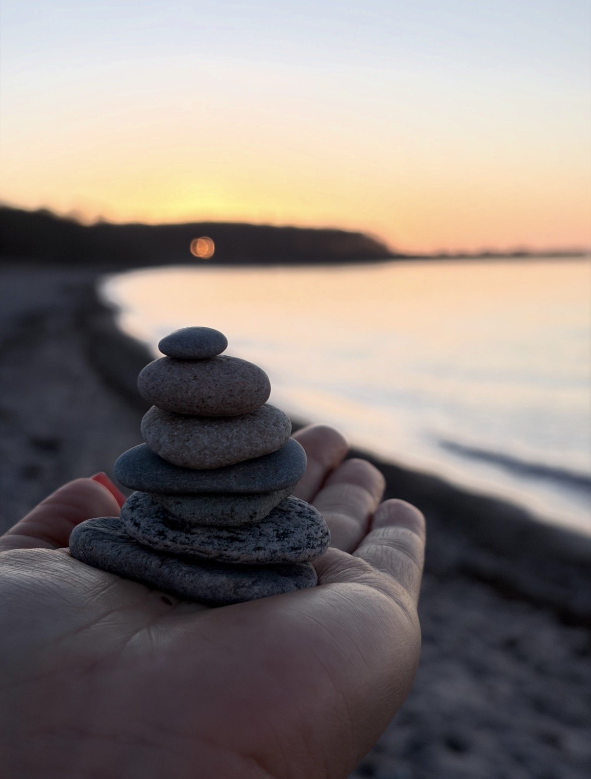 Eine Hand hält einen Stapel von glatten Steinen vor einem Strand bei Sonnenuntergang, mit Wasser und einem bewölkten Himmel im Hintergrund.