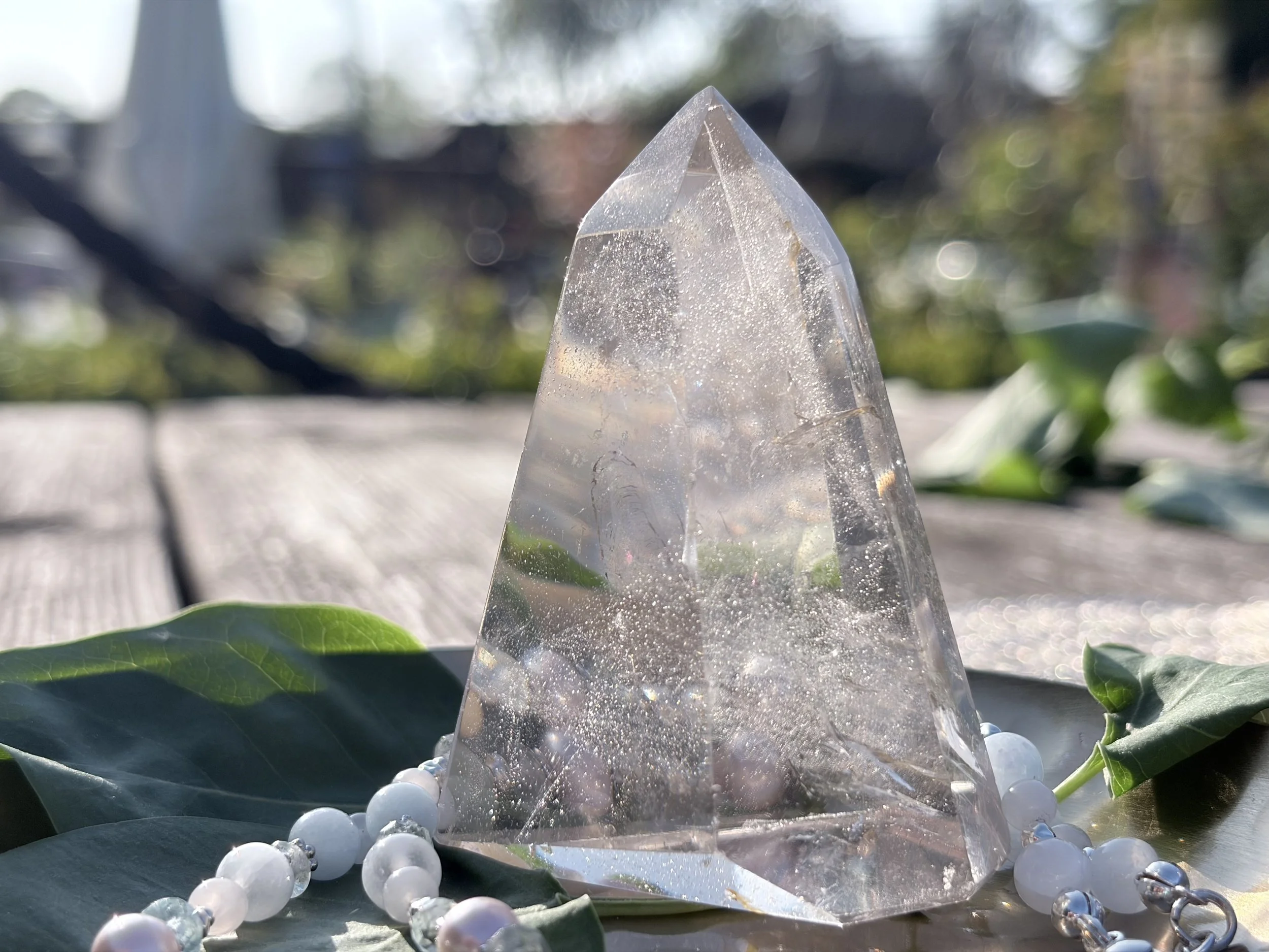 Ein transparenter, keilförmiger Kristall, umgeben von weißen Perlen auf grünen Blättern bei Sonnenlicht.