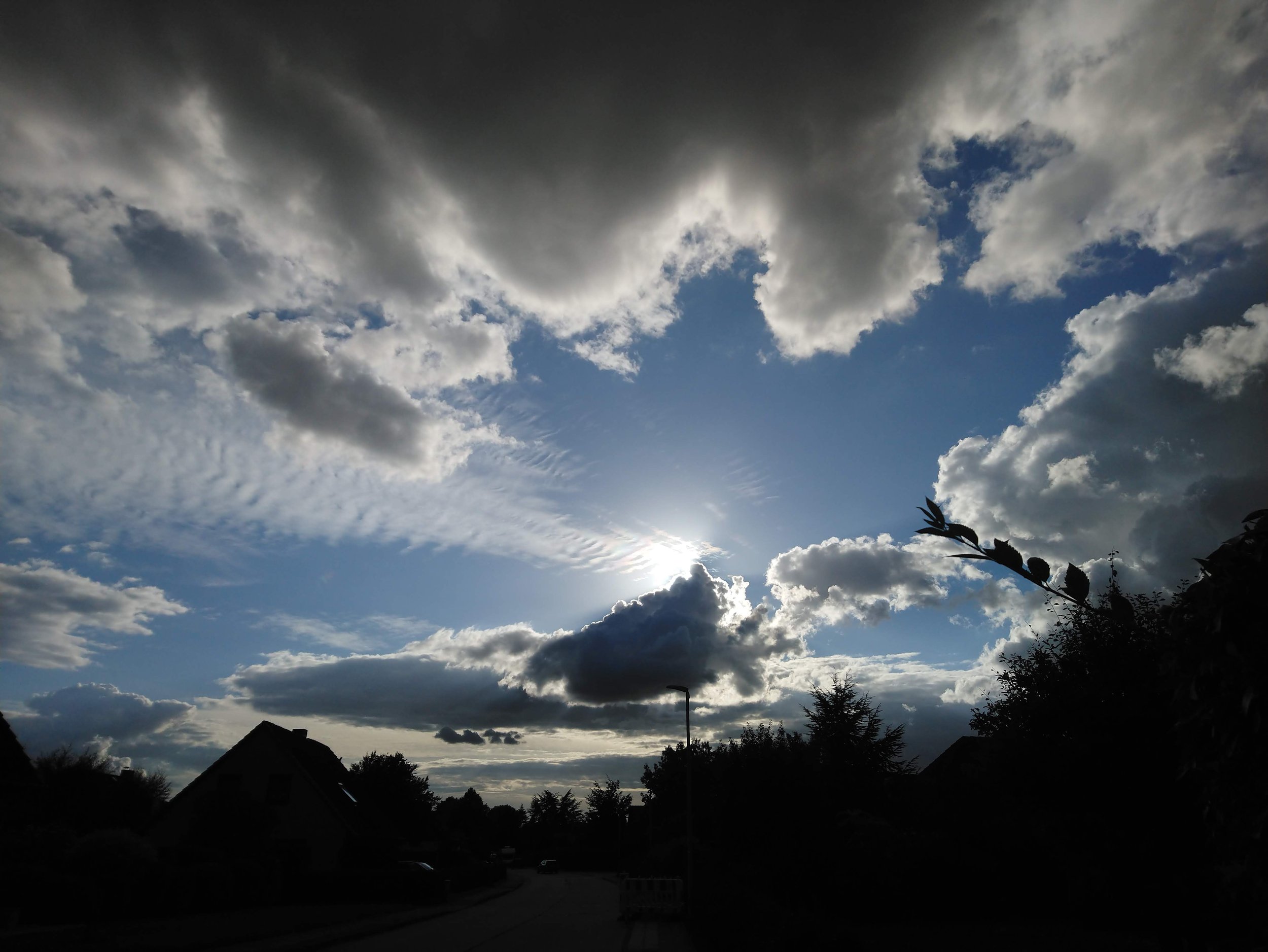 Der Himmel ist teilweise bewölkt mit dunklen und hellen Wolken, und Sonnenlicht scheint hinter den Wolken hindurch. Im Vordergrund sind Silhouetten von Häusern, Bäumen und einem Laternenmast sichtbar.