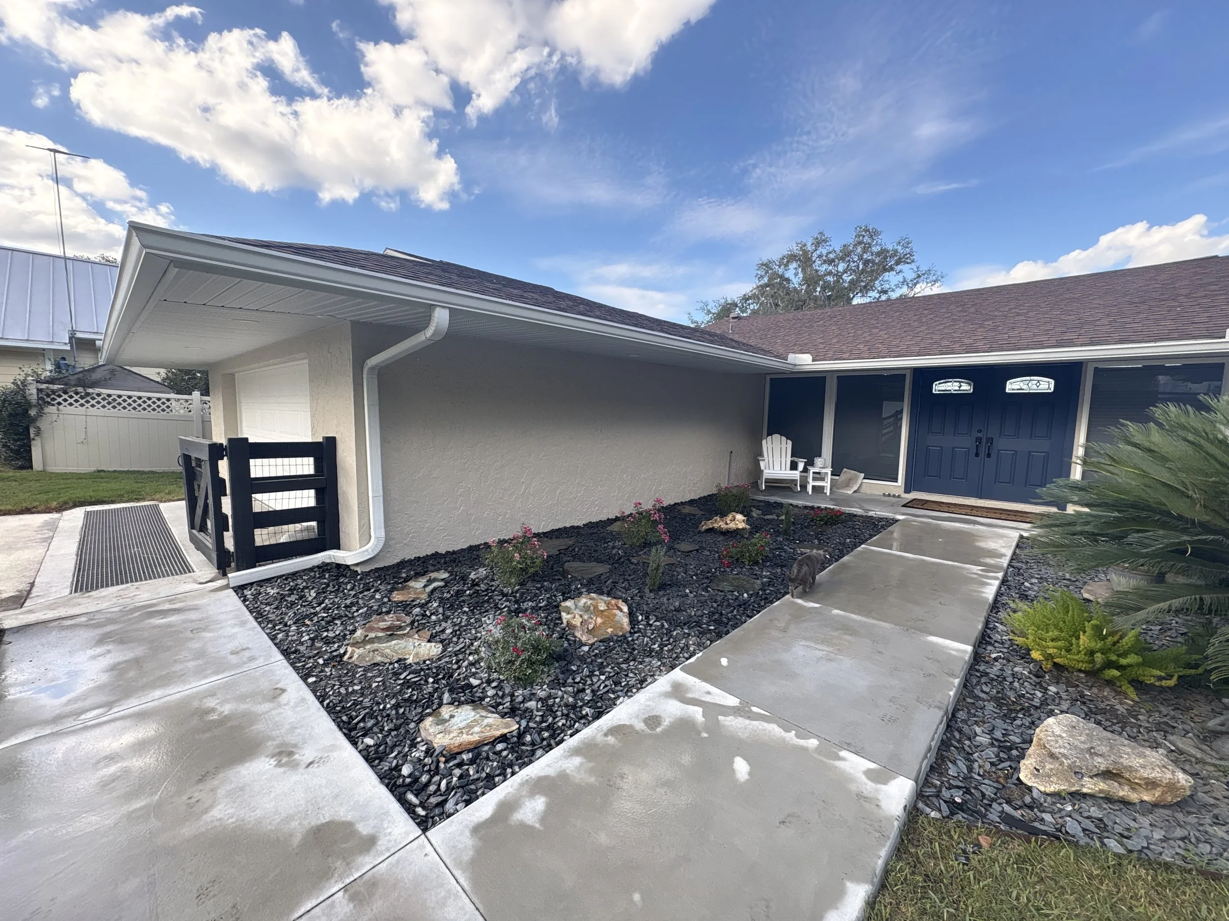 A house with a garage, dark blue front door, and small front garden with plants, rocks, and a concrete walkway under a partly cloudy sky.