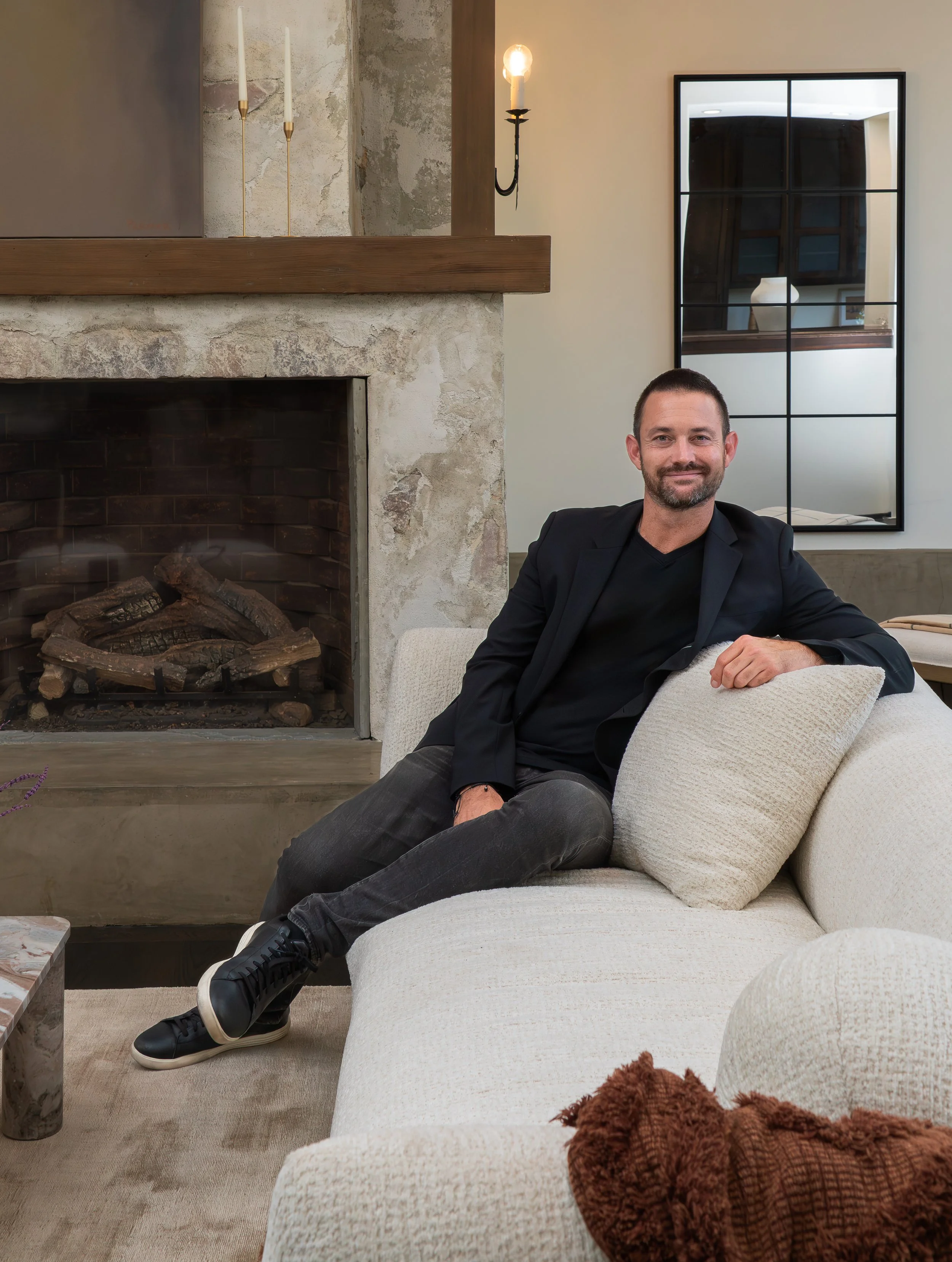 A man with short dark hair and a beard sitting on a cream-colored sofa, smiling, wearing a black blazer and dark gray pants, holding a beige pillow, in a living room with a fireplace and a large mirror.