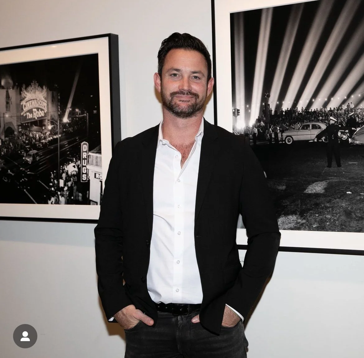 A man with dark hair and a beard, wearing a black blazer and white shirt, standing in front of black-and-white framed photographs at an art gallery or exhibition.