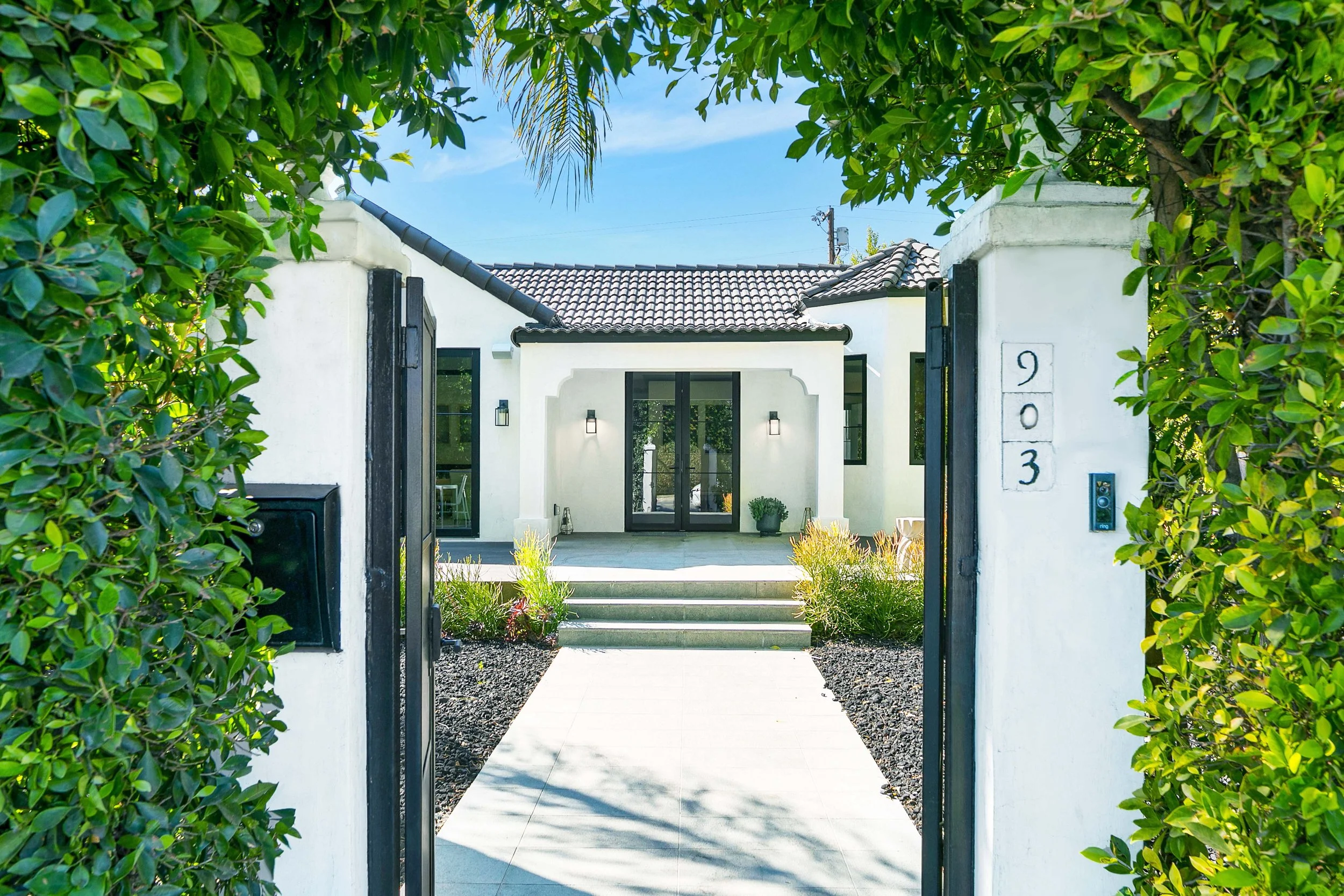 View through an open black gate showing a modern white house with a tiled roof, small front yard, and potted plant, framed by lush green bushes and trees.