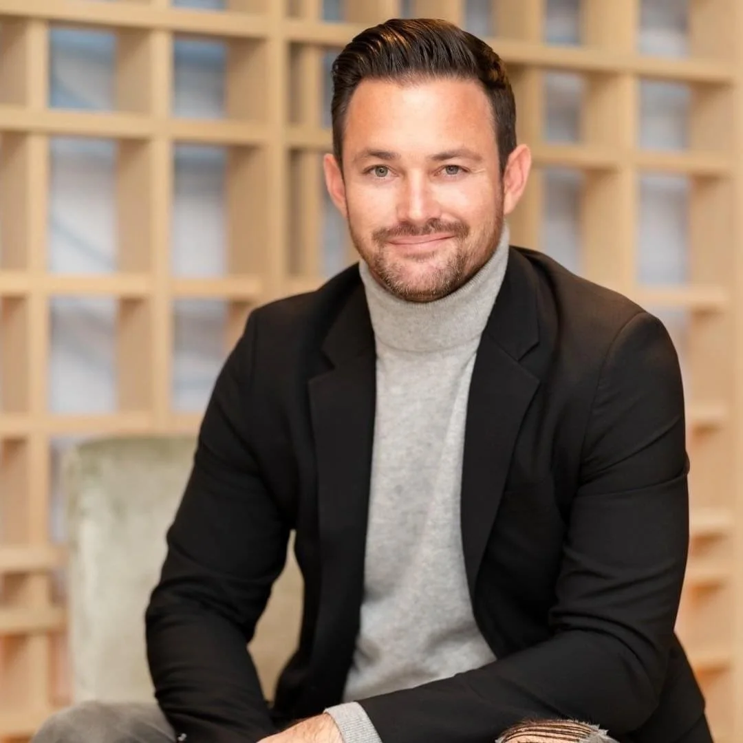 A man with dark hair and a beard, smiling, wearing a gray turtleneck sweater and a black blazer, sitting in front of a wooden lattice background.