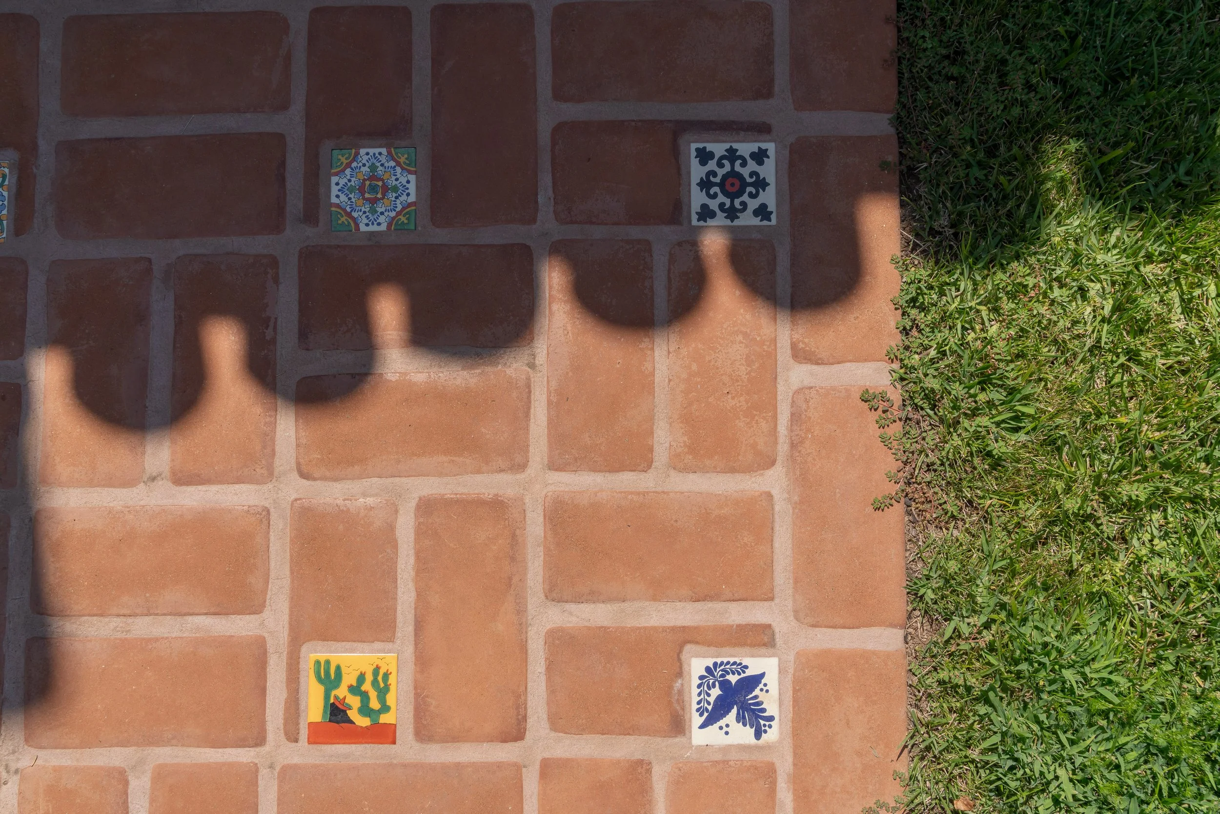 Colorful decorative tiles on a brick pathway with grass on the right side. Shadows of two people cast onto the tiles. Three tiles are visible, each with different designs: one with a floral pattern, another with a geometric pattern, and the third with a cactus and bird motif.