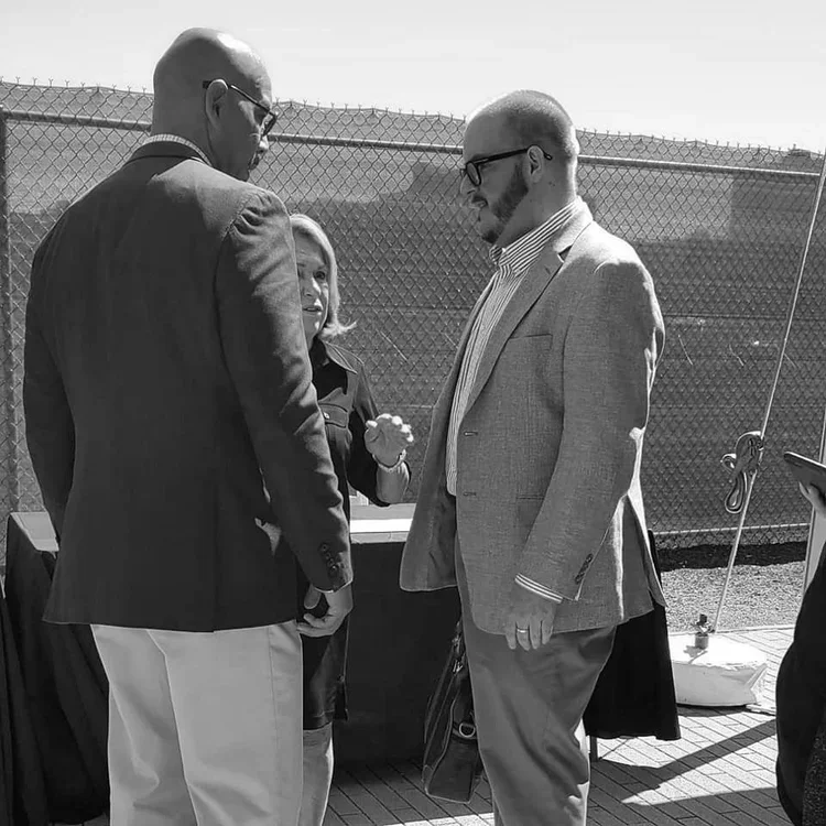 Three people having a conversation outdoors, two men and one woman, with a chain-link fence in the background.