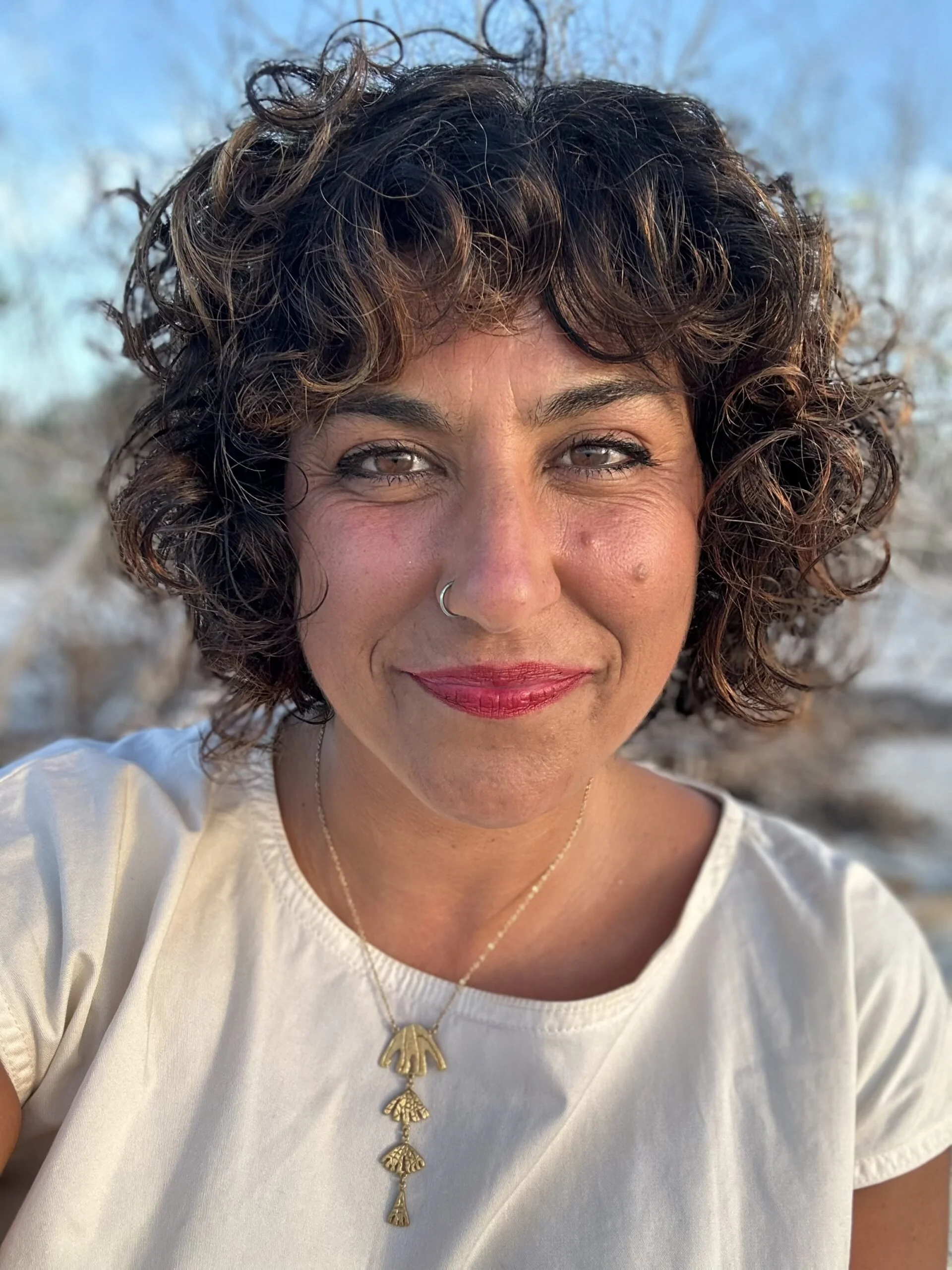 A close-up of a woman with curly brown hair, wearing a white shirt, a gold necklace with multiple leaf-shaped pendants, and a nose ring, outdoors with a blue sky and trees in the background.