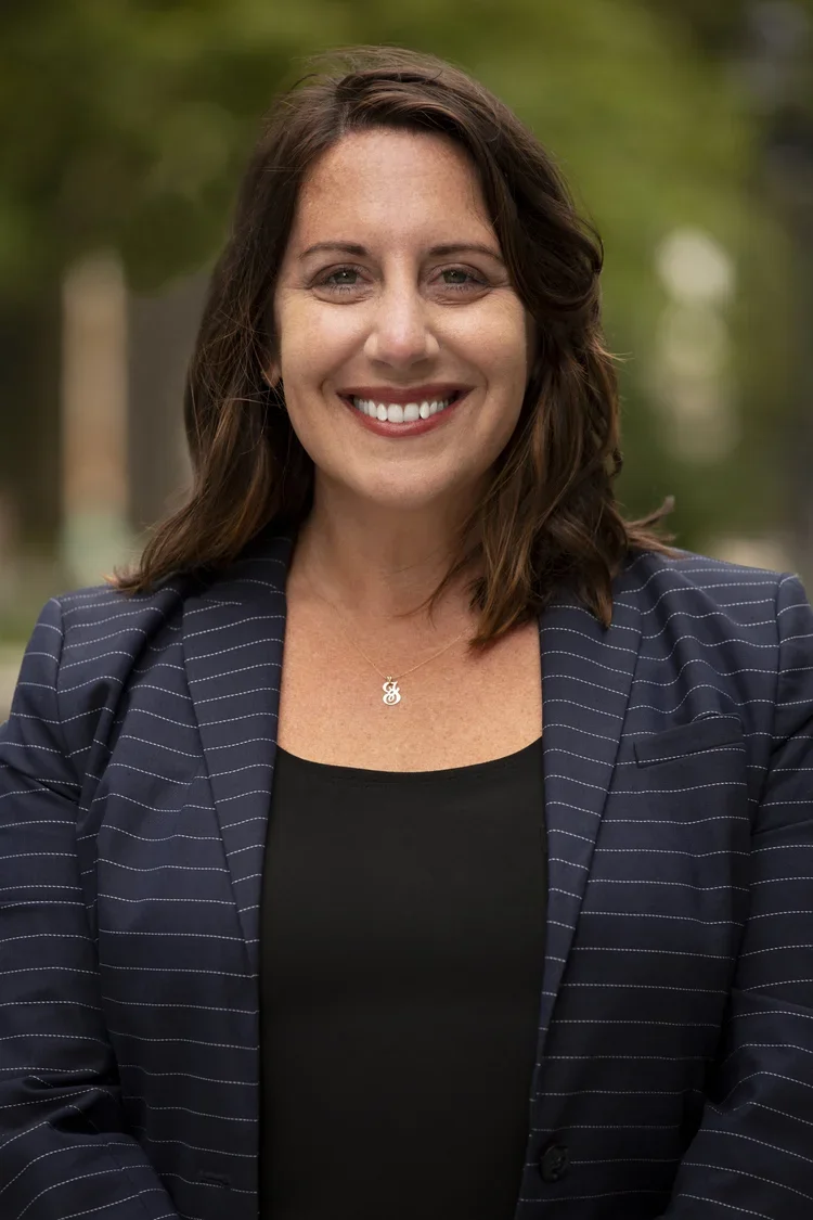 A woman with shoulder-length brown hair smiling, wearing a black top, navy blazer with thin white stripes, and a necklace with a charm, standing outdoors with green foliage in the background.