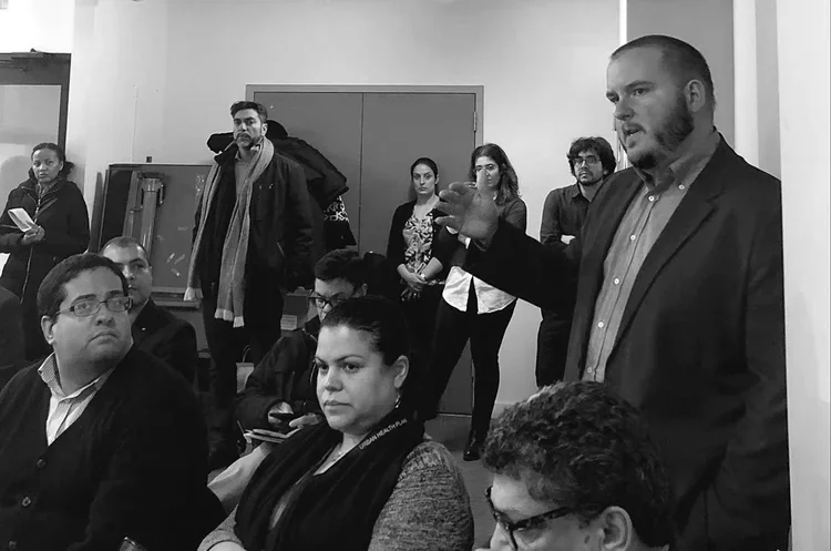 A man in a blazer and shirt is speaking and gesturing with his hand to a group of seated and standing people in a room, engaging in a discussion or presentation.