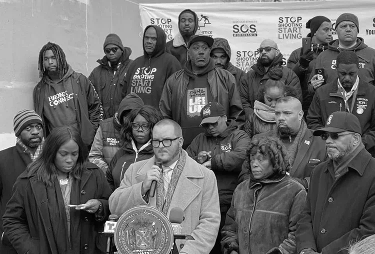 A group of people gathered outdoors, with some wearing jackets and hats, listening to Michael Brady speaking into a microphone at a public event advocating for stopping gun violence and promoting social justice.