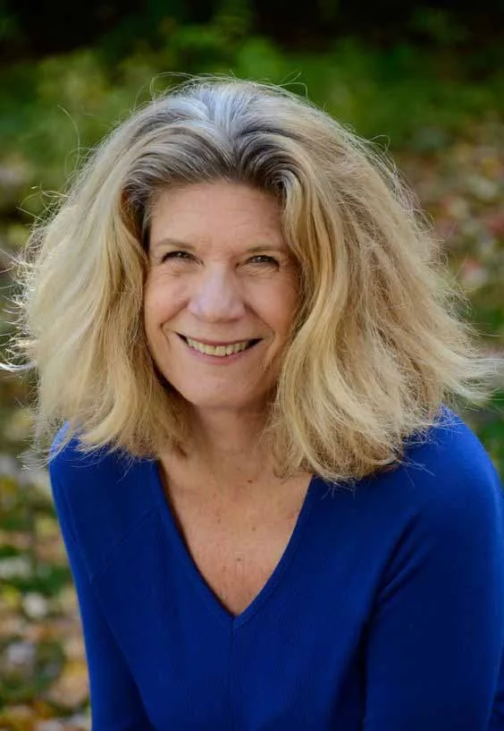 Smiling middle-aged woman with shoulder-length blonde hair outdoors, wearing a blue top, with green foliage in the background.