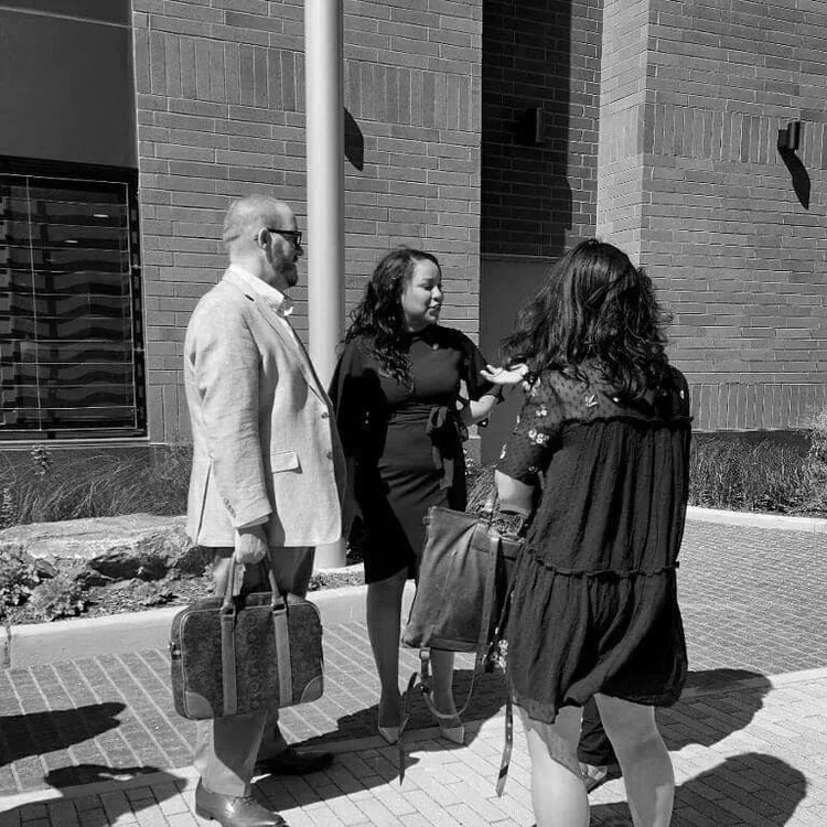 Three people standing on a sidewalk outside a brick building, engaged in conversation. One man carries a briefcase, and two women hold bags.