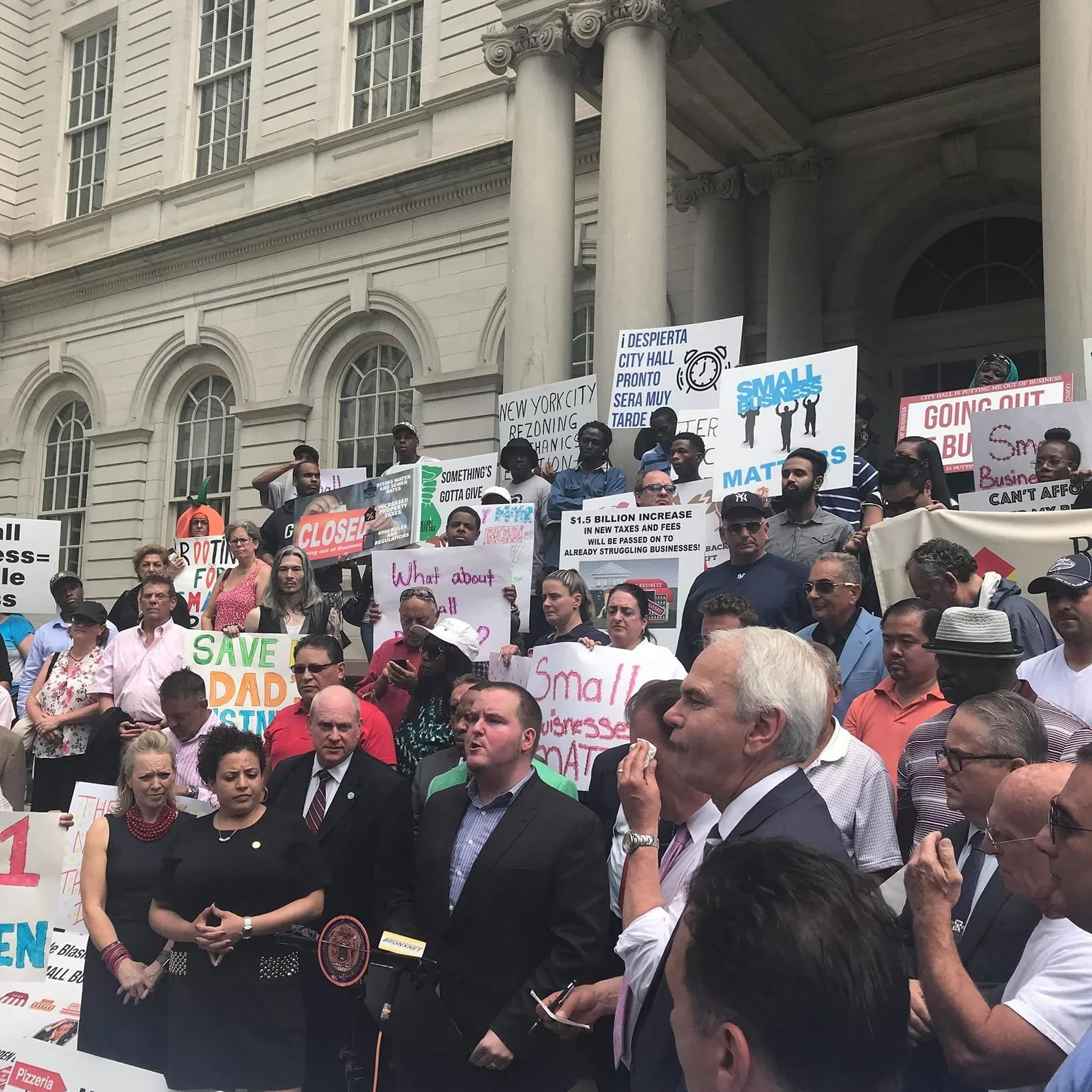 A crowd of people protesting outside a government building with signs and banners advocating for small businesses and against taxes and fees.